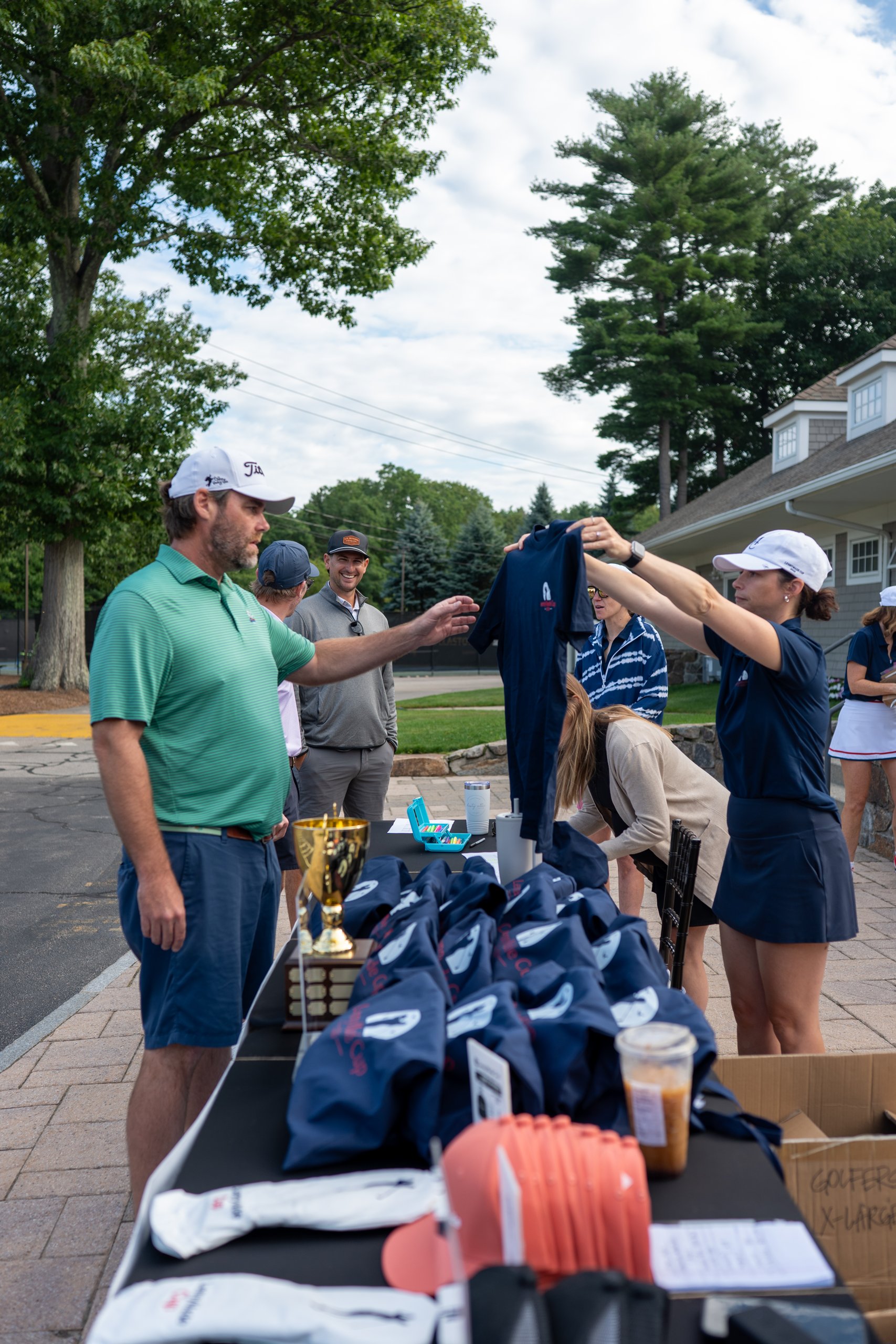 Registration staff holding up a navy LennyMade Cup t-shirt for a golfer at check-in, the trophy and gift bags on the table.