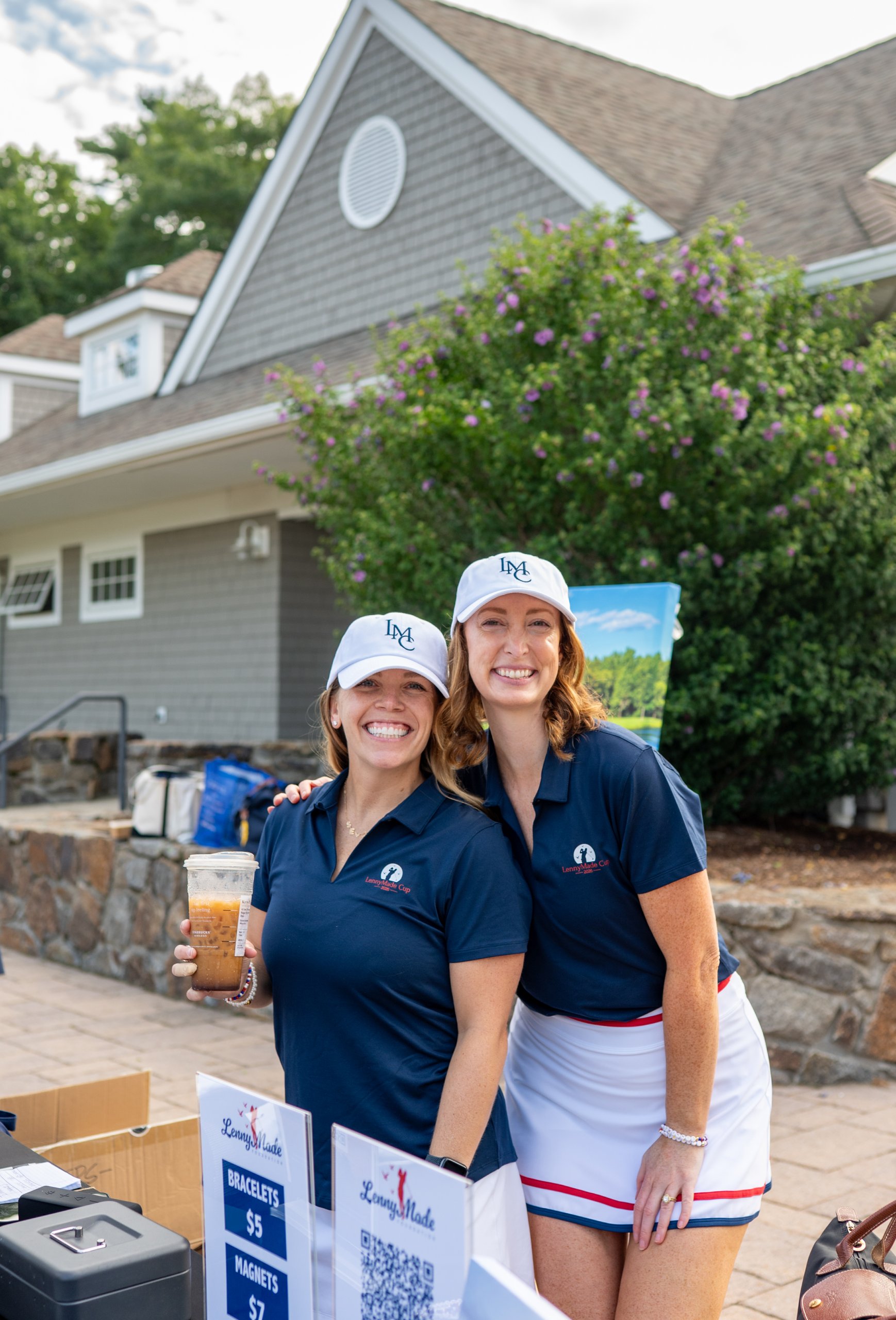 Two volunteers in LMC caps and navy LennyMade Cup polos smiling behind the merchandise table, one holding an iced coffee.