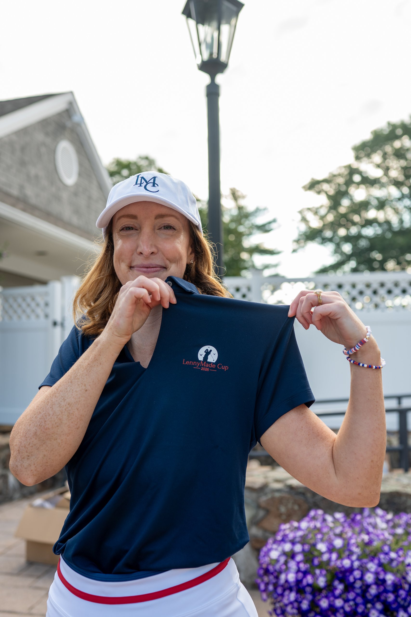 Volunteer in an LMC cap holding up a navy LennyMade Cup 2026 polo shirt on the clubhouse patio.