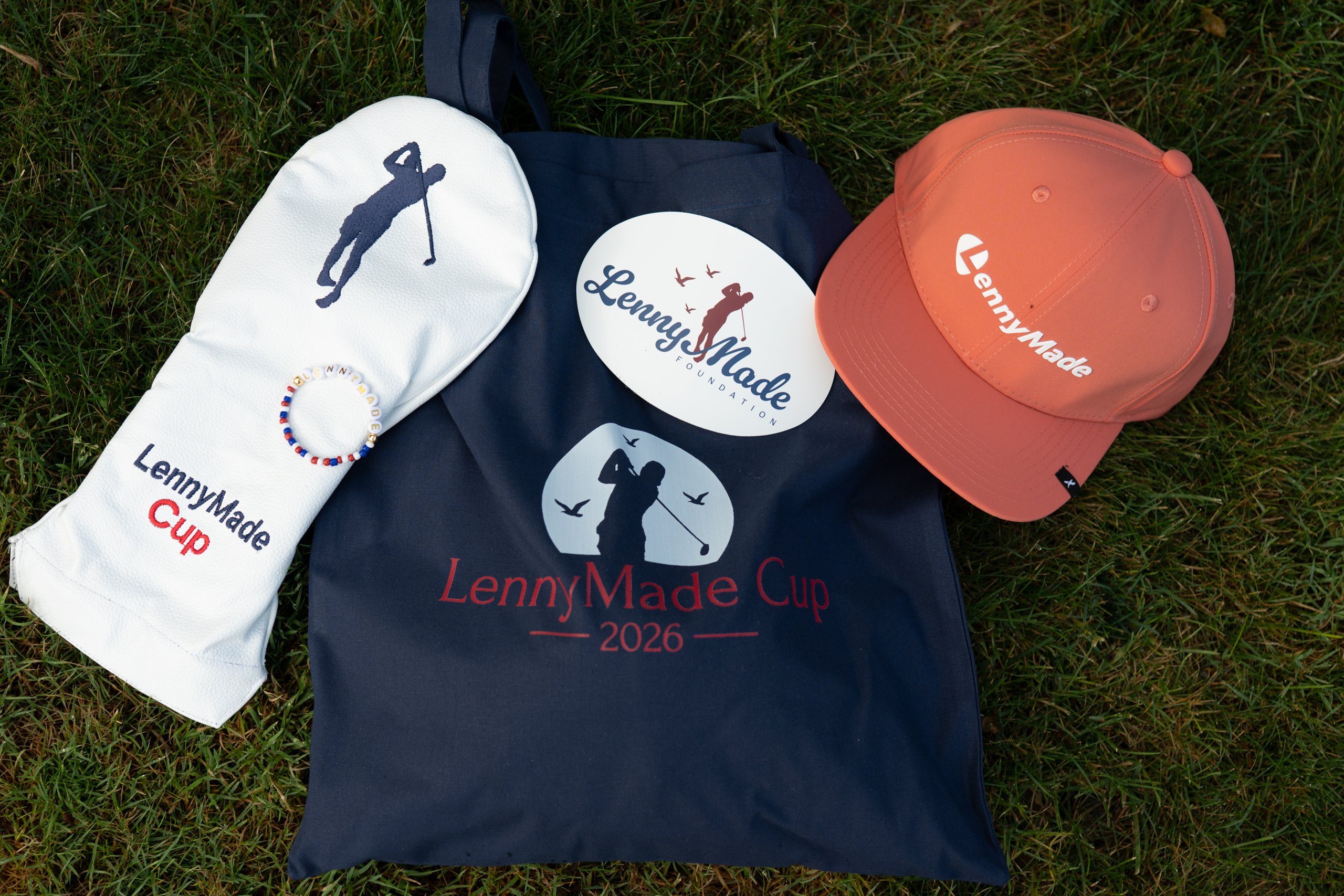 Flat lay on the grass of LennyMade Cup swag: a white driver headcover, navy LennyMade Cup 2026 tote, foundation sticker and a coral LennyMade cap.