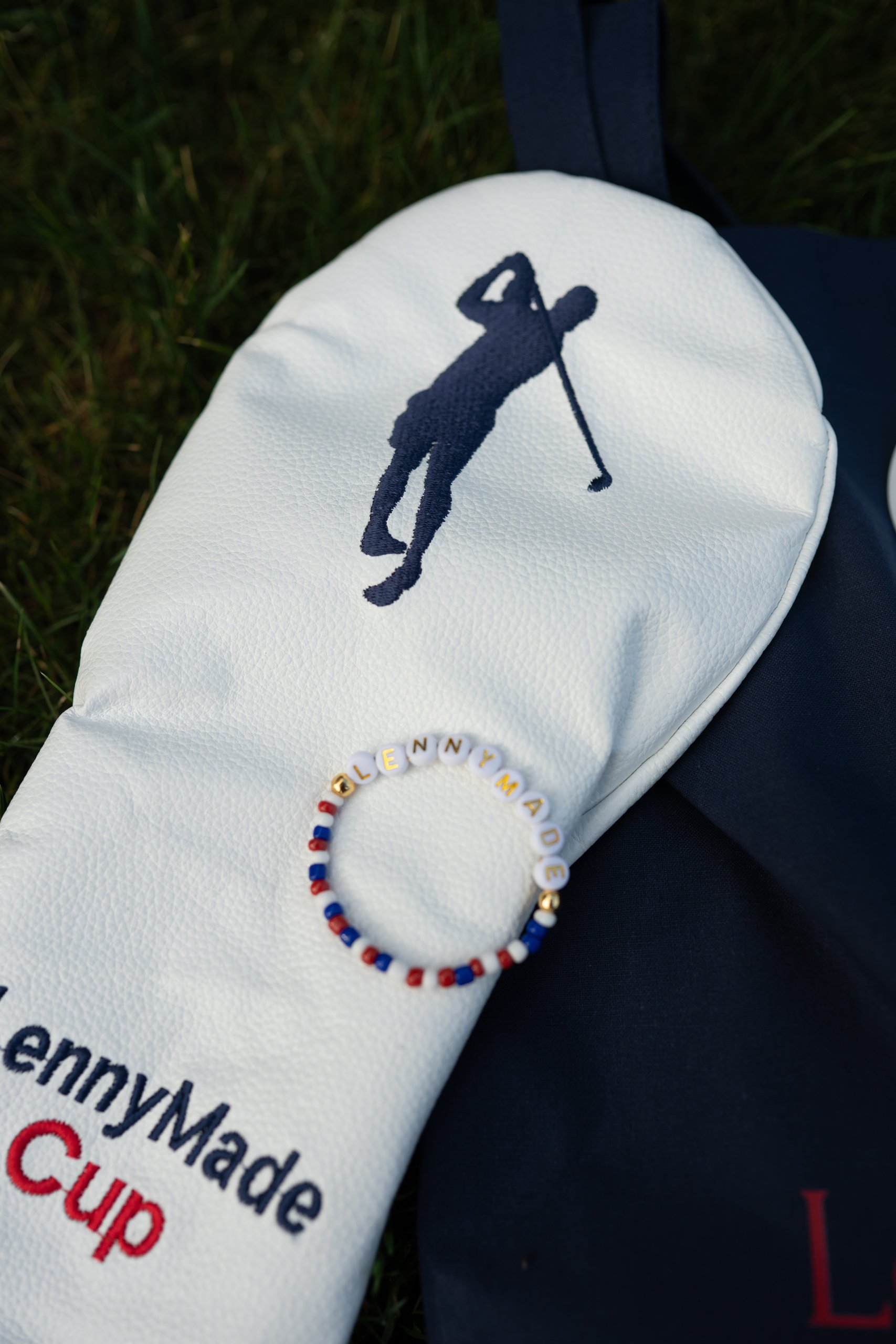 Close-up flat lay of a white LennyMade Cup driver headcover with a beaded LENNYMADE bracelet resting on a navy tote.