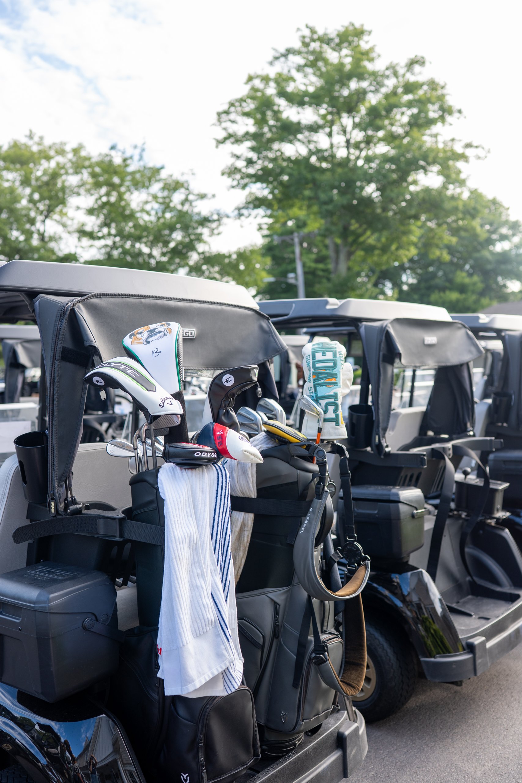 Row of golf carts staged with players' bags and clubs, including novelty driver headcovers, under the trees.