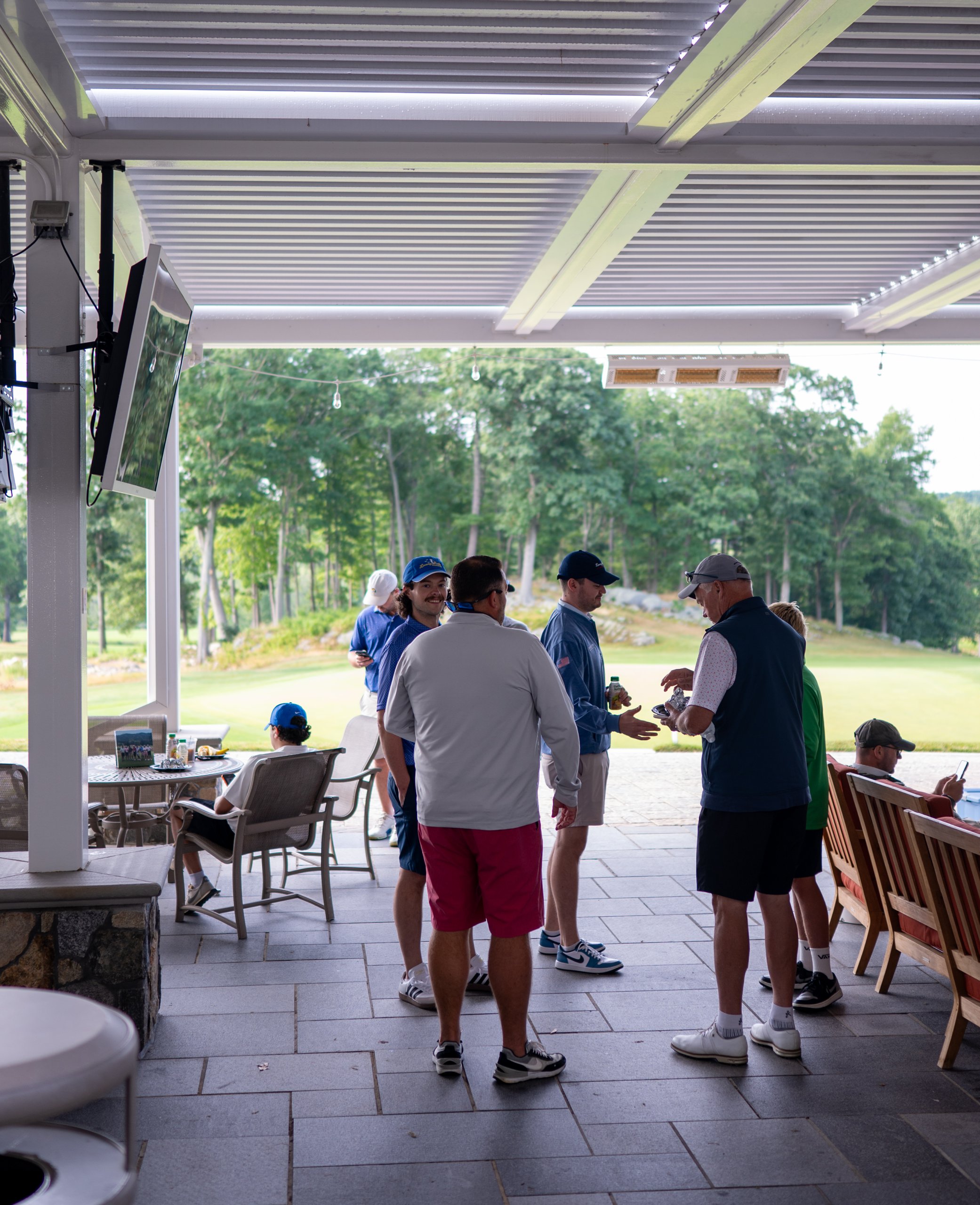 Golfers mingling on the covered clubhouse patio before the round, the course visible beyond.