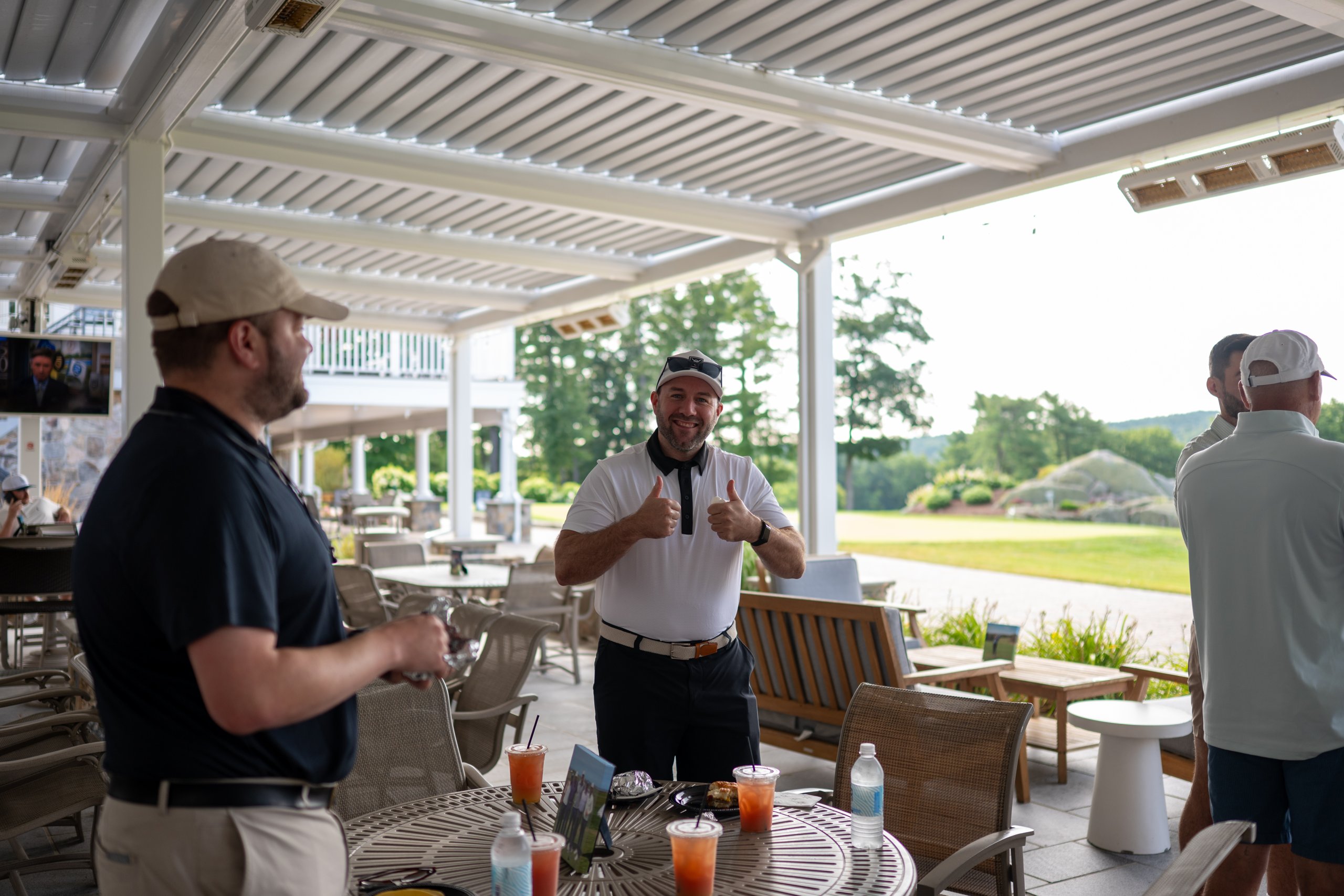 A golfer giving two thumbs up on the clubhouse patio as others chat over drinks.