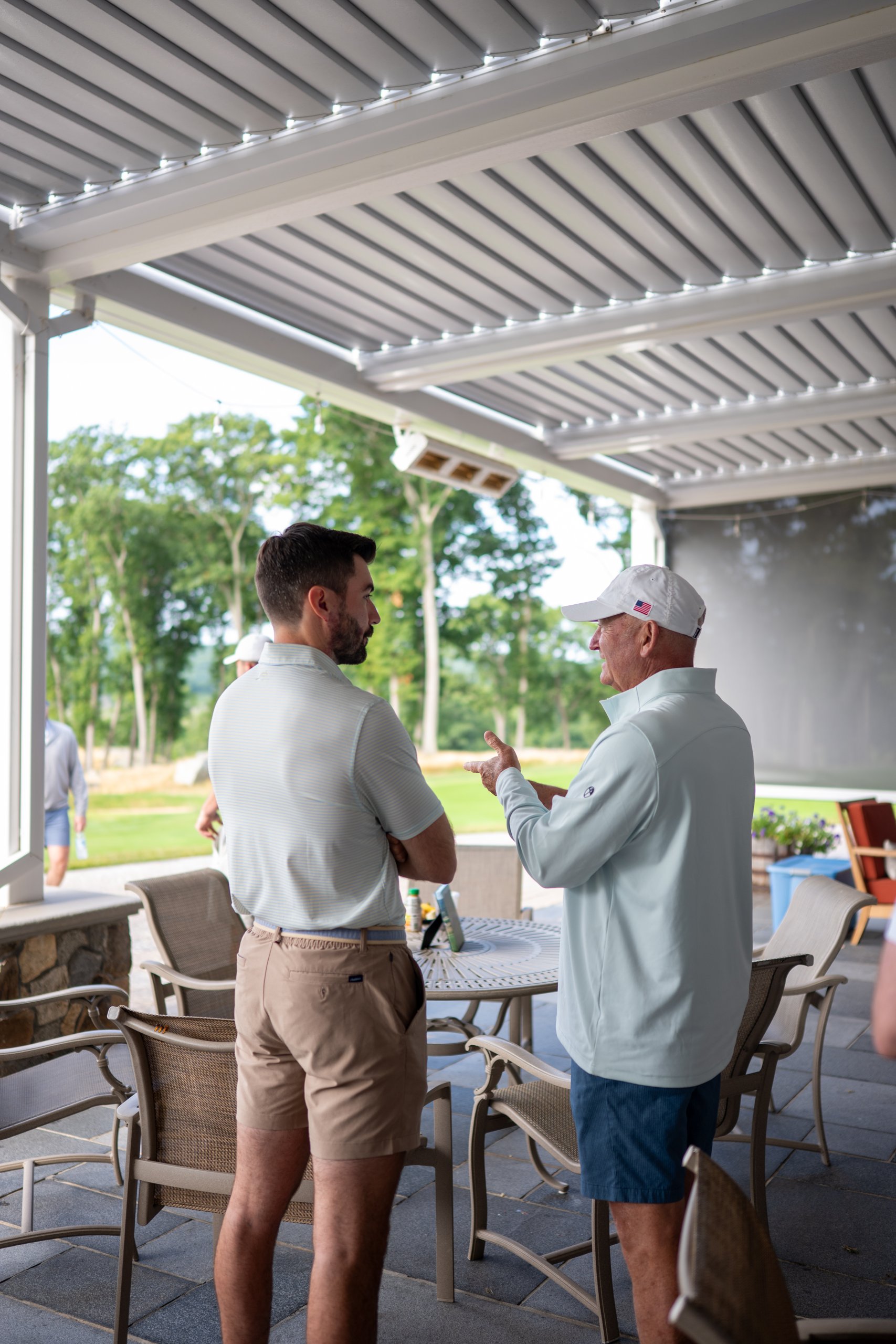Two golfers talking on the patio, an older man in a US-flag cap gesturing, the fairway behind them.