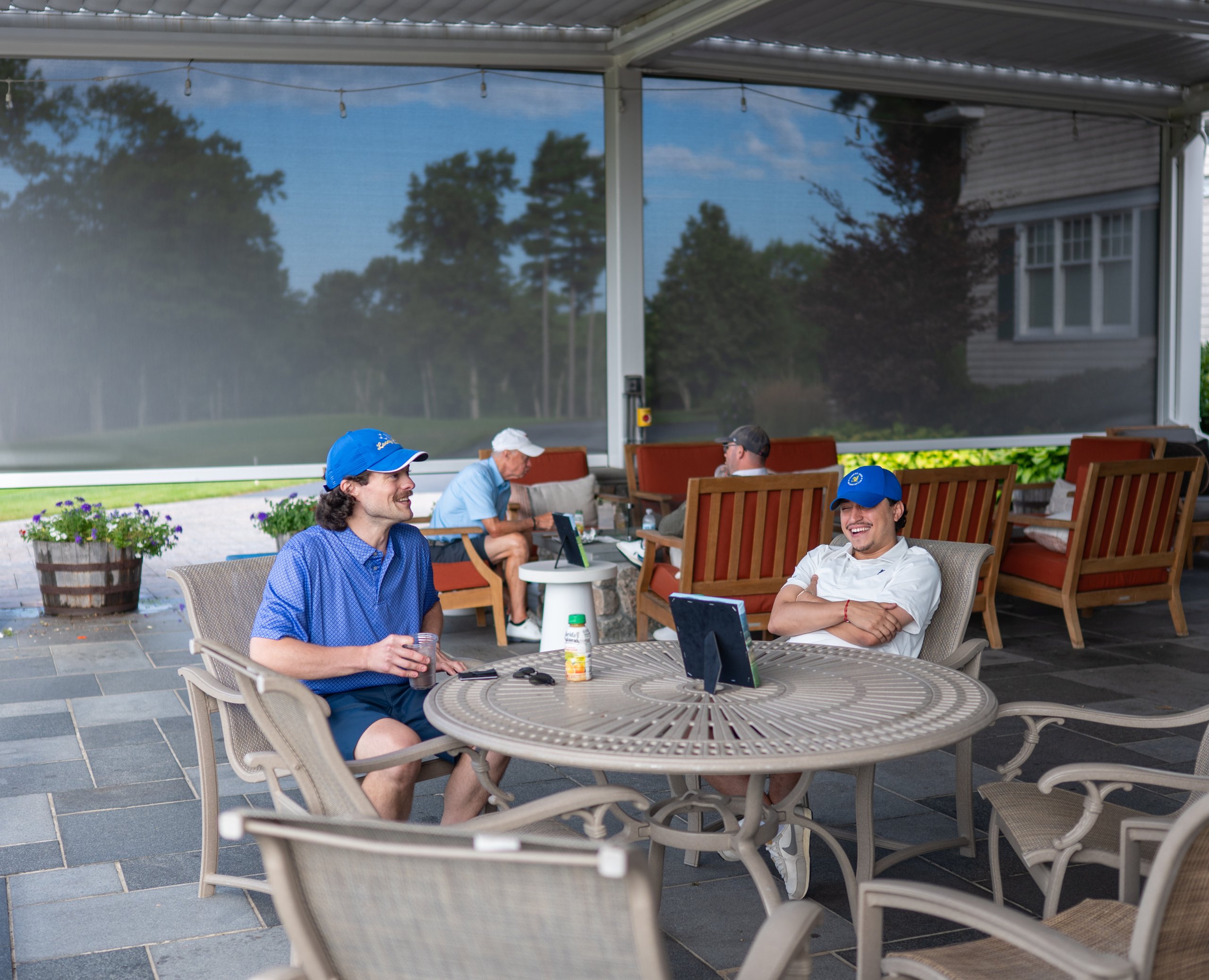 Two golfers laughing together at a patio table under the pergola, the course visible through the shade screen behind.