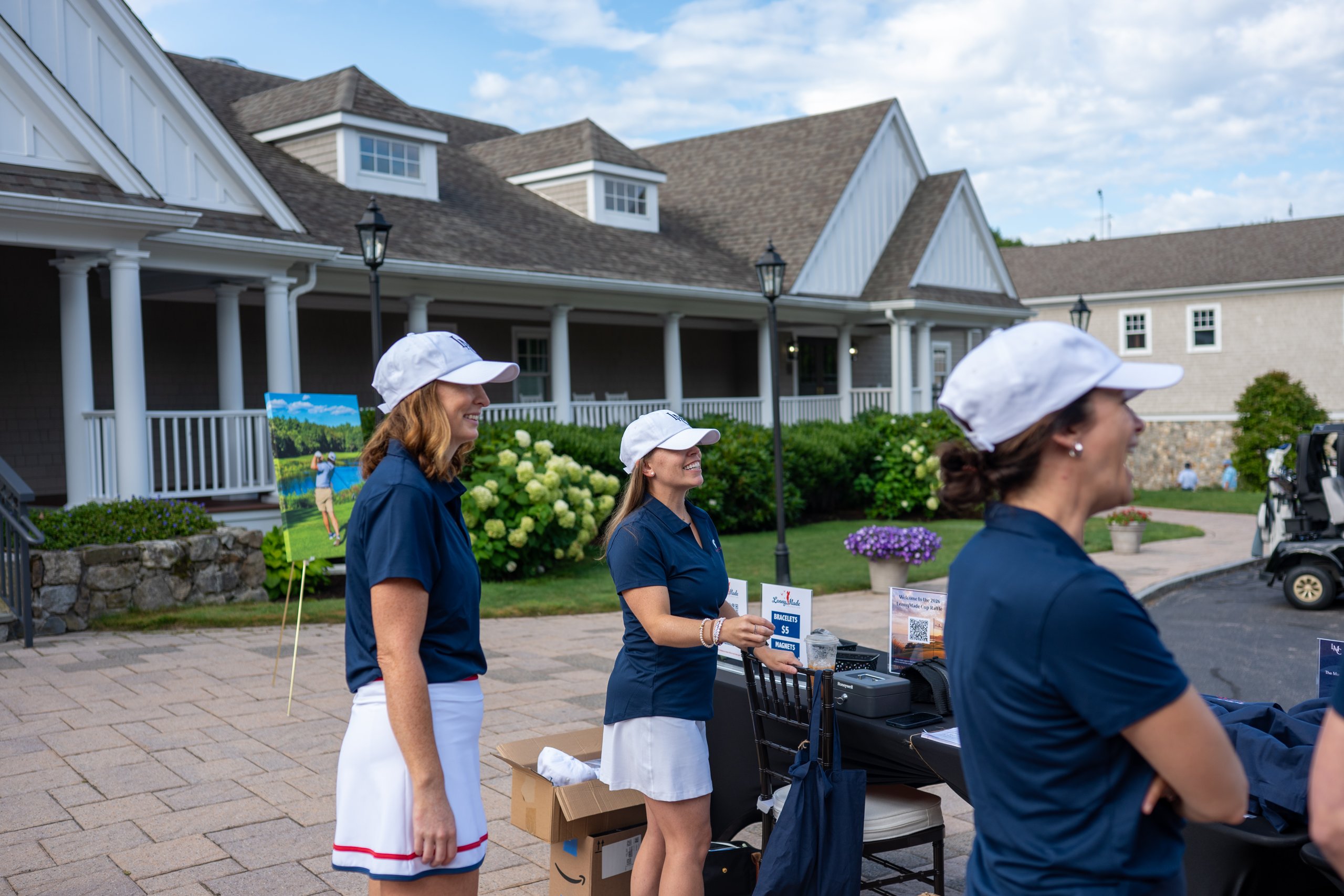 Three LennyMade Cup volunteers in LMC caps and navy polos at the registration table in front of the clubhouse.