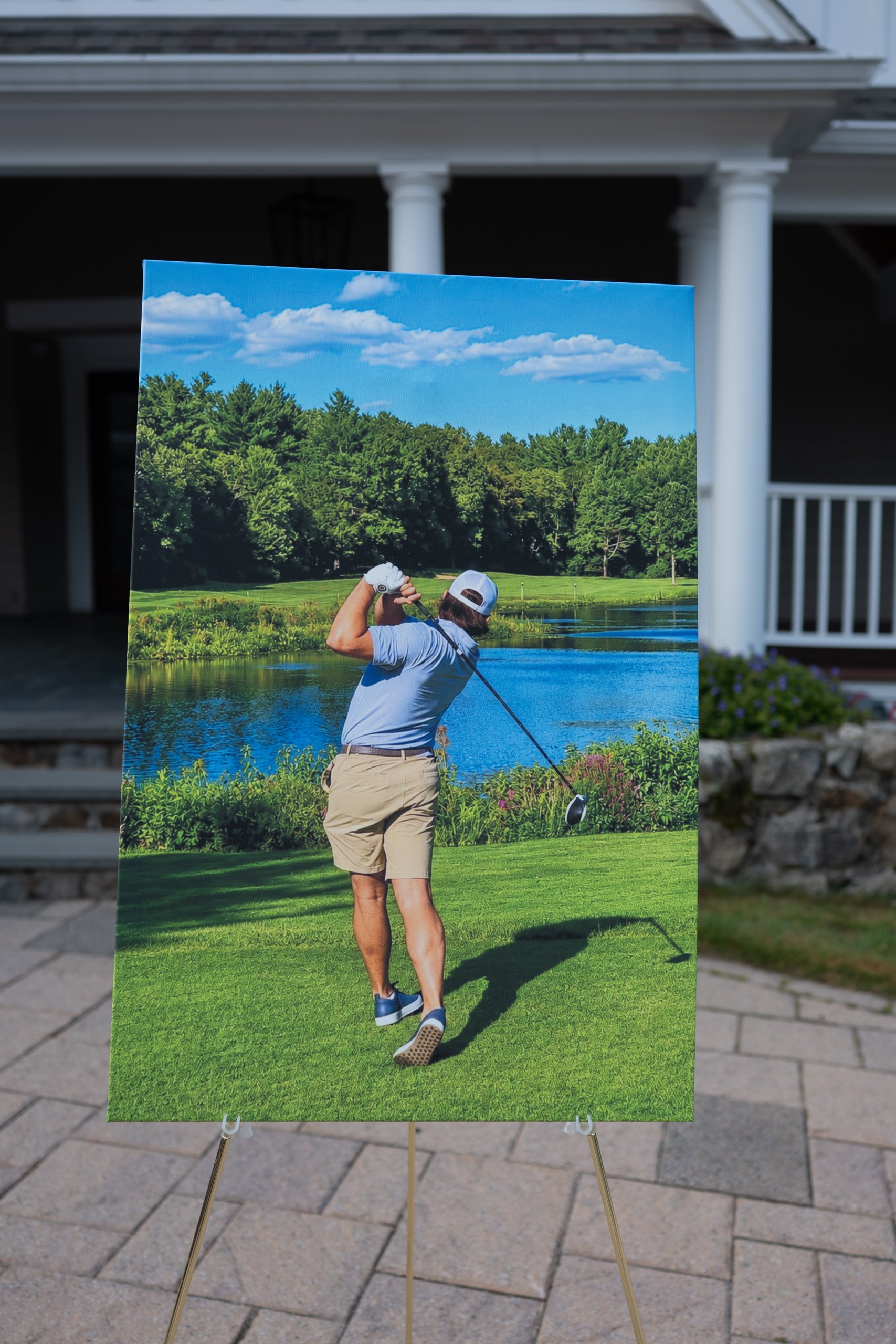 A large canvas print of a golfer teeing off toward a pond, displayed on an easel in front of the clubhouse.
