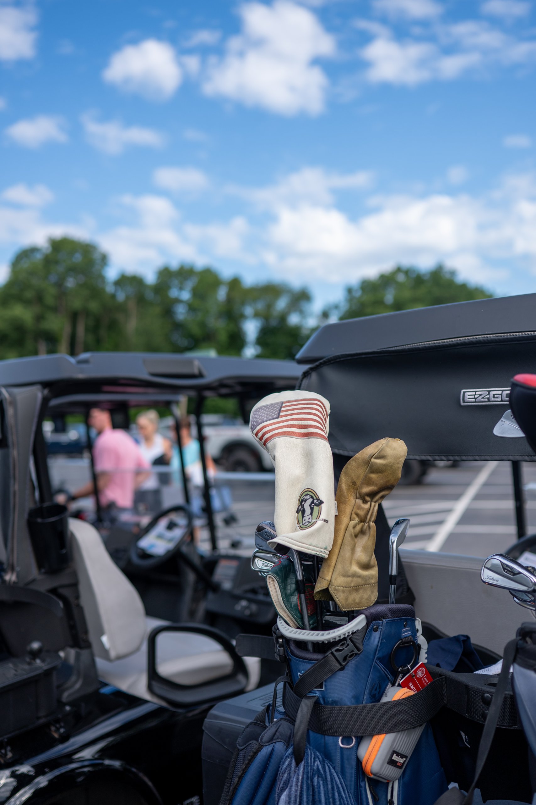 Golf bags loaded on carts with a US-flag driver headcover and a cow-emblem headcover, players gathering behind.