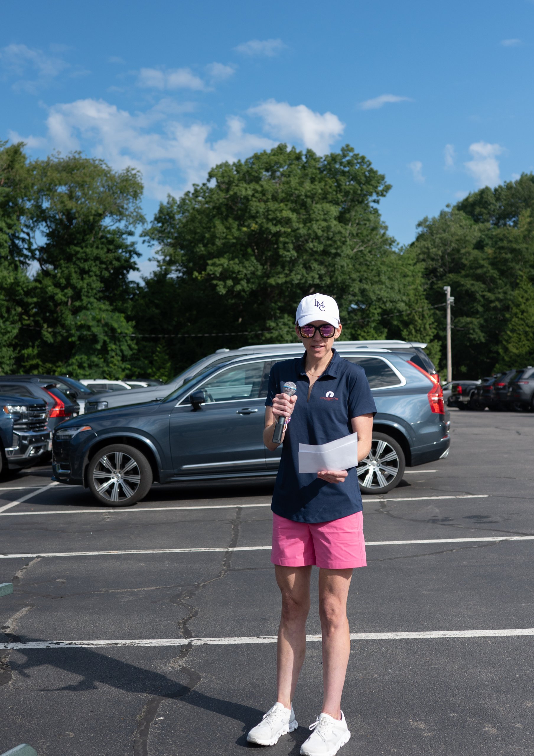 Event host in an LMC cap, navy polo and pink shorts reading announcements into a microphone in the parking lot.