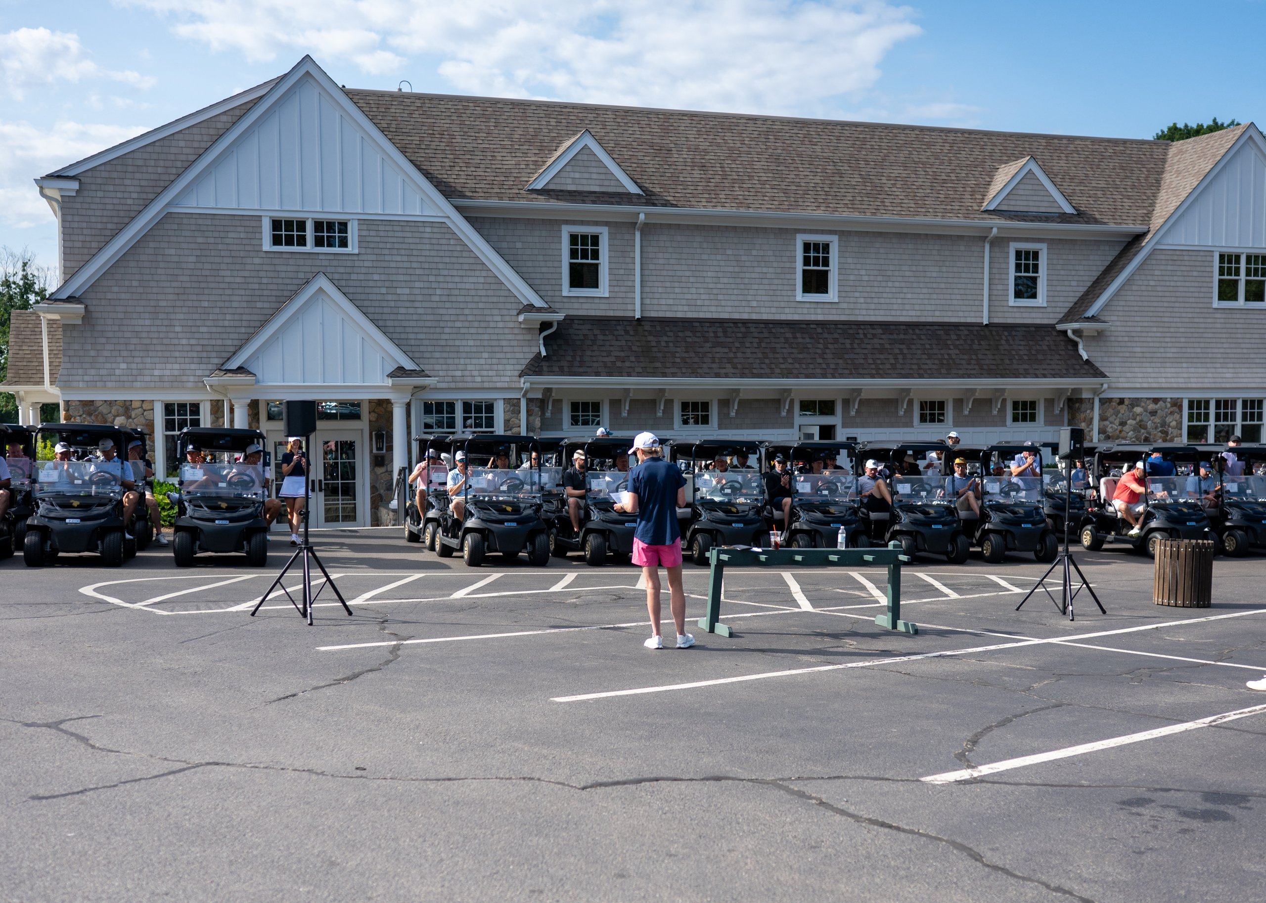Wide view of the shotgun-start gathering, rows of black golf carts lined up in front of the clubhouse as the host addresses players.