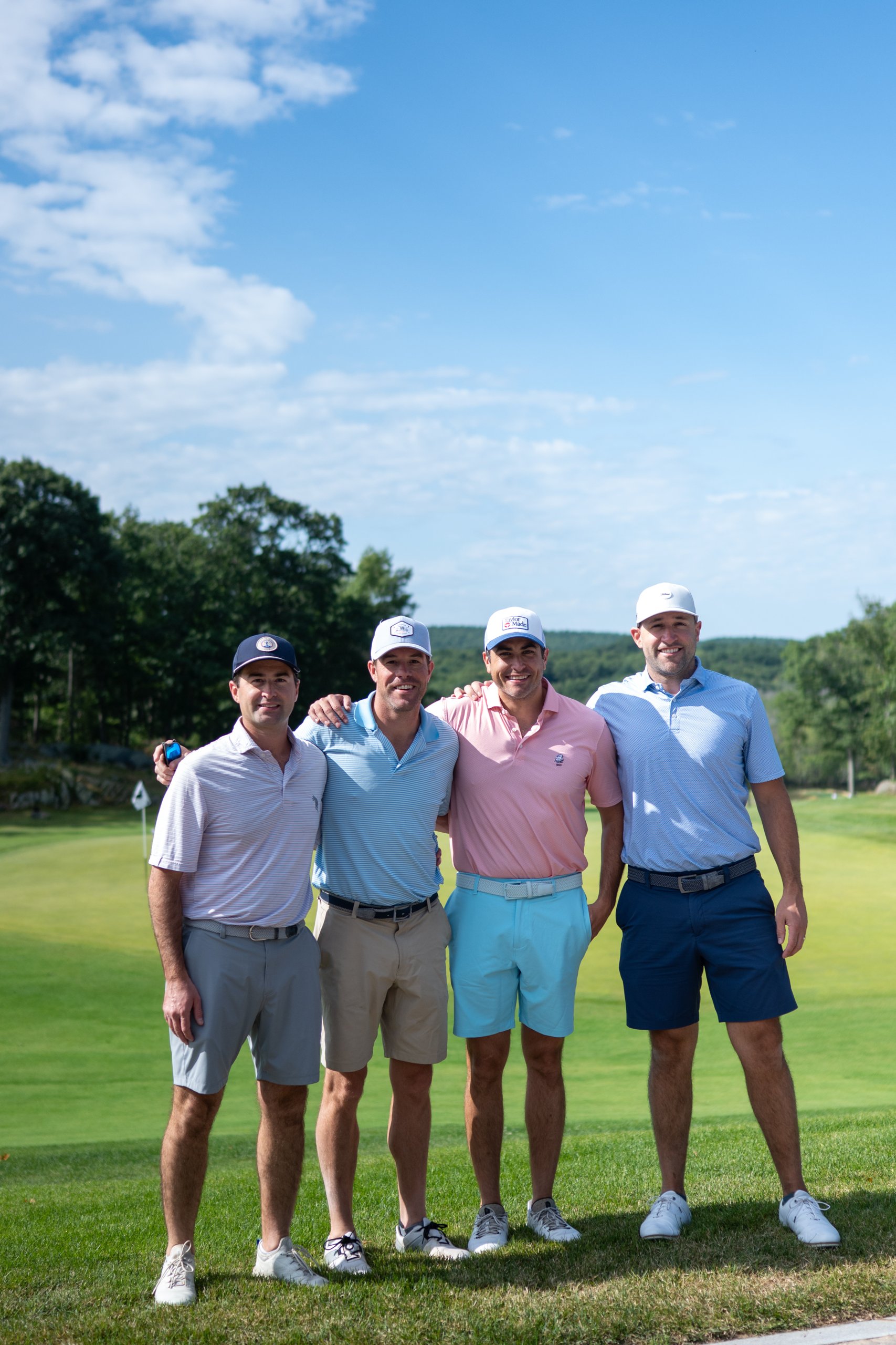 Foursome of four men posing arm in arm on the course, wooded hills behind them.