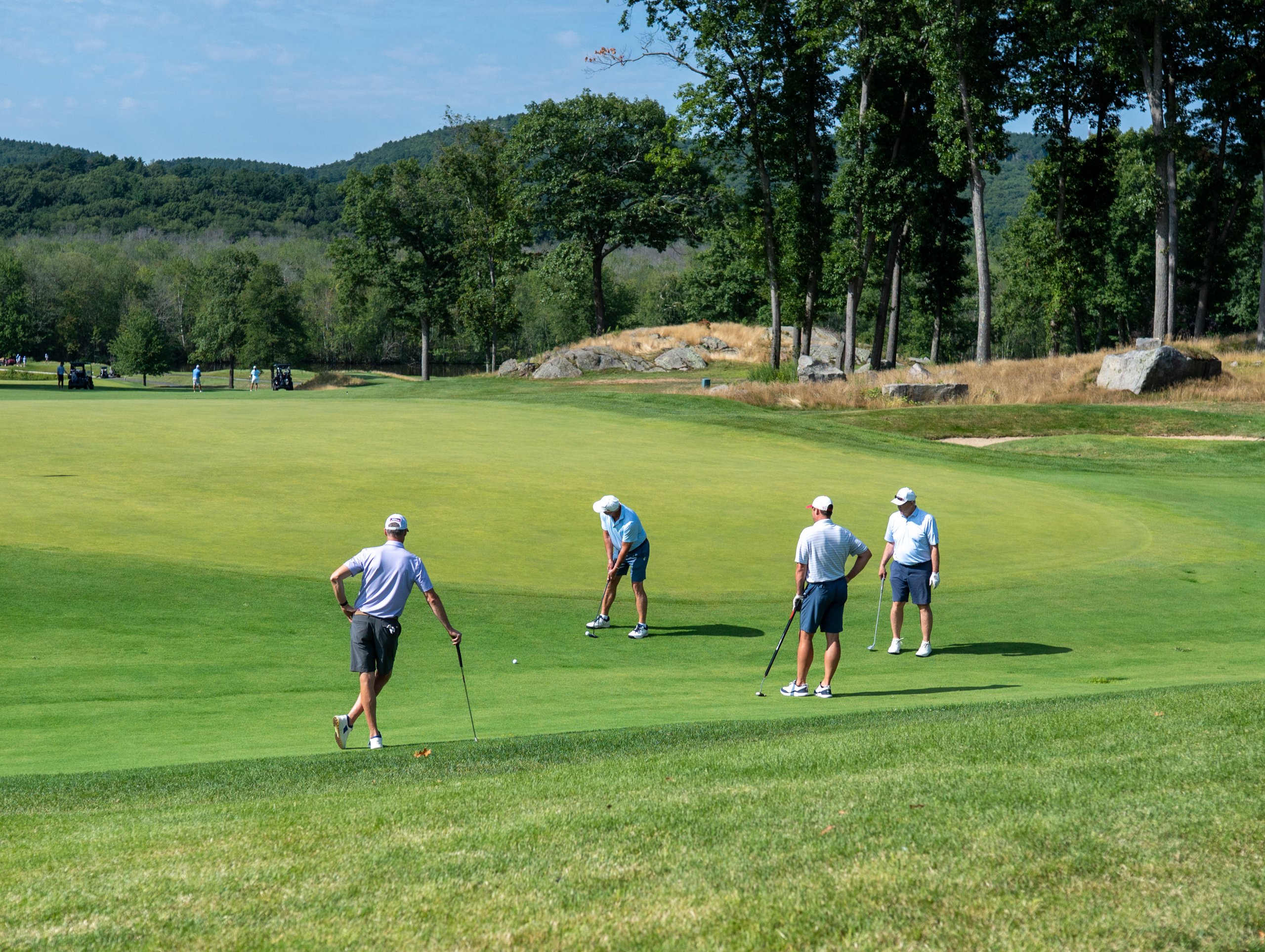 Foursome on the green, one lining up a putt while the others watch, a rocky outcrop and hills behind.