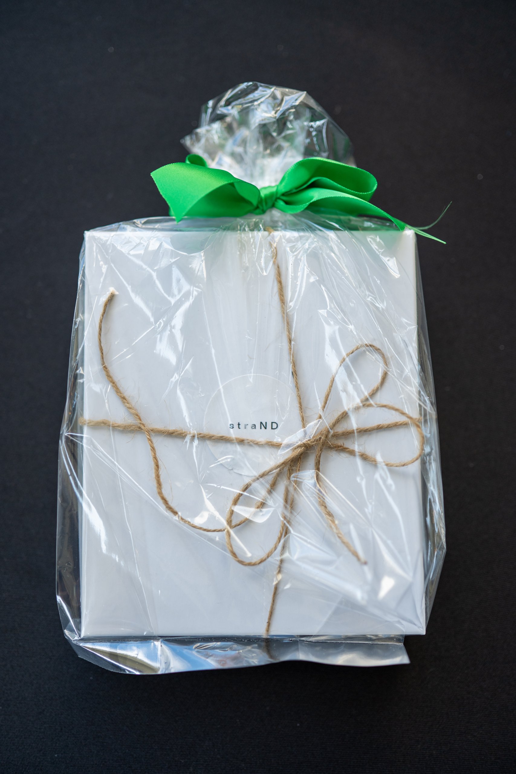 A white straND gift box wrapped in cellophane with twine and a green bow on a black tablecloth.