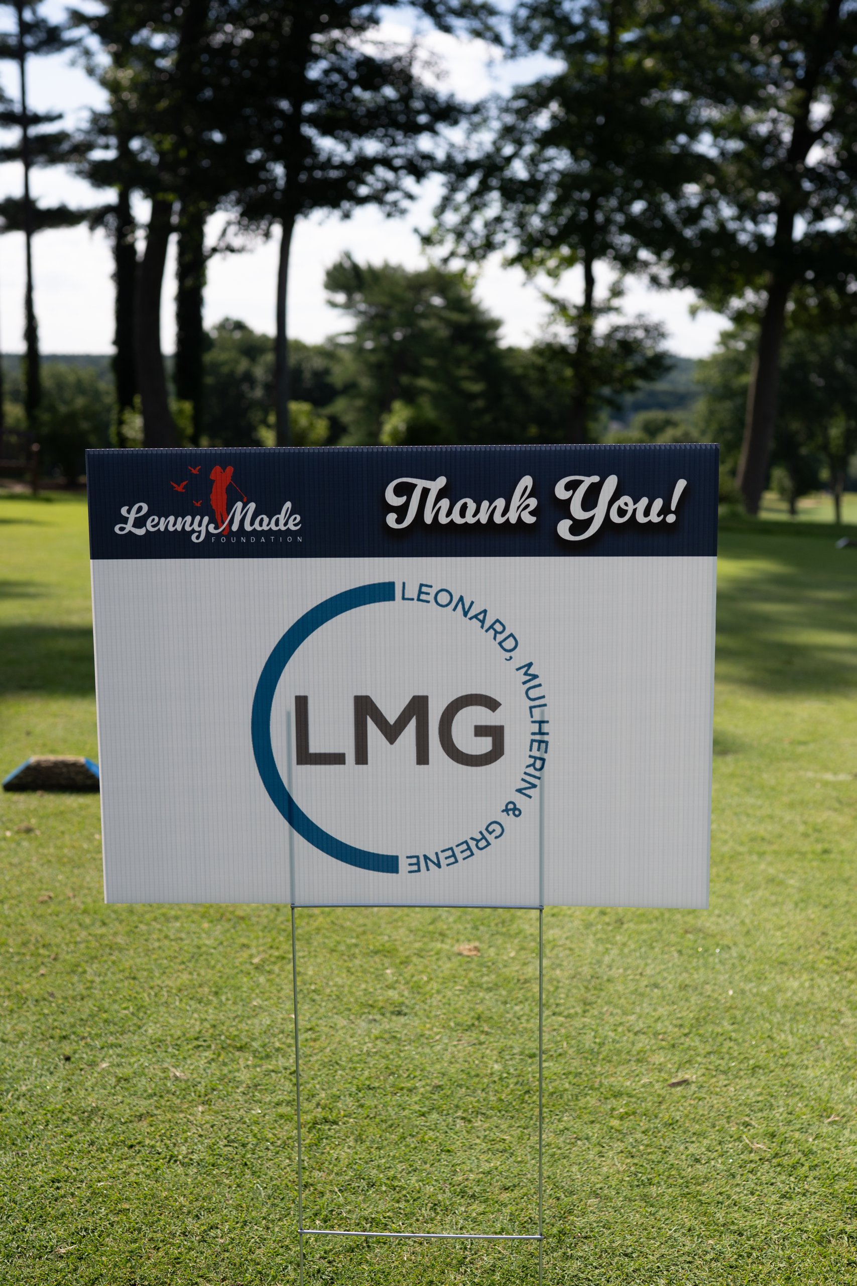 A LennyMade Foundation Thank You sponsor sign for LMG, Leonard, Mulherin and Greene, on the fairway.