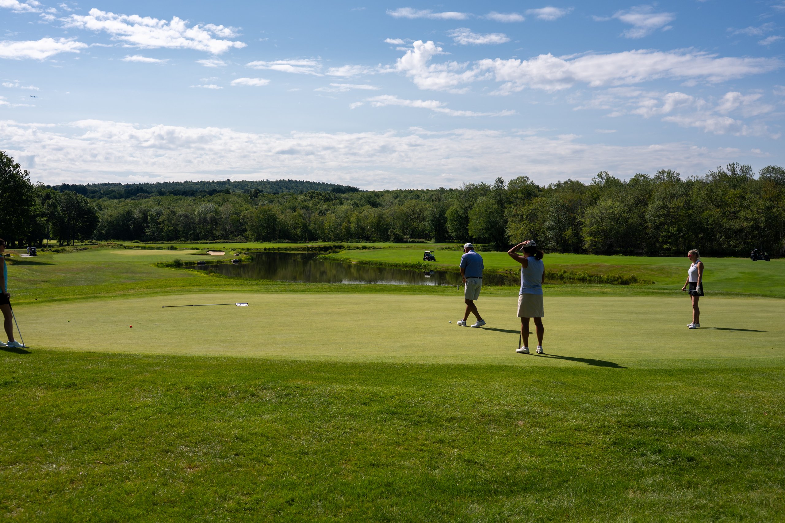 Wide view of a foursome on the green reading their putts, a pond and dense treeline stretching across the background.