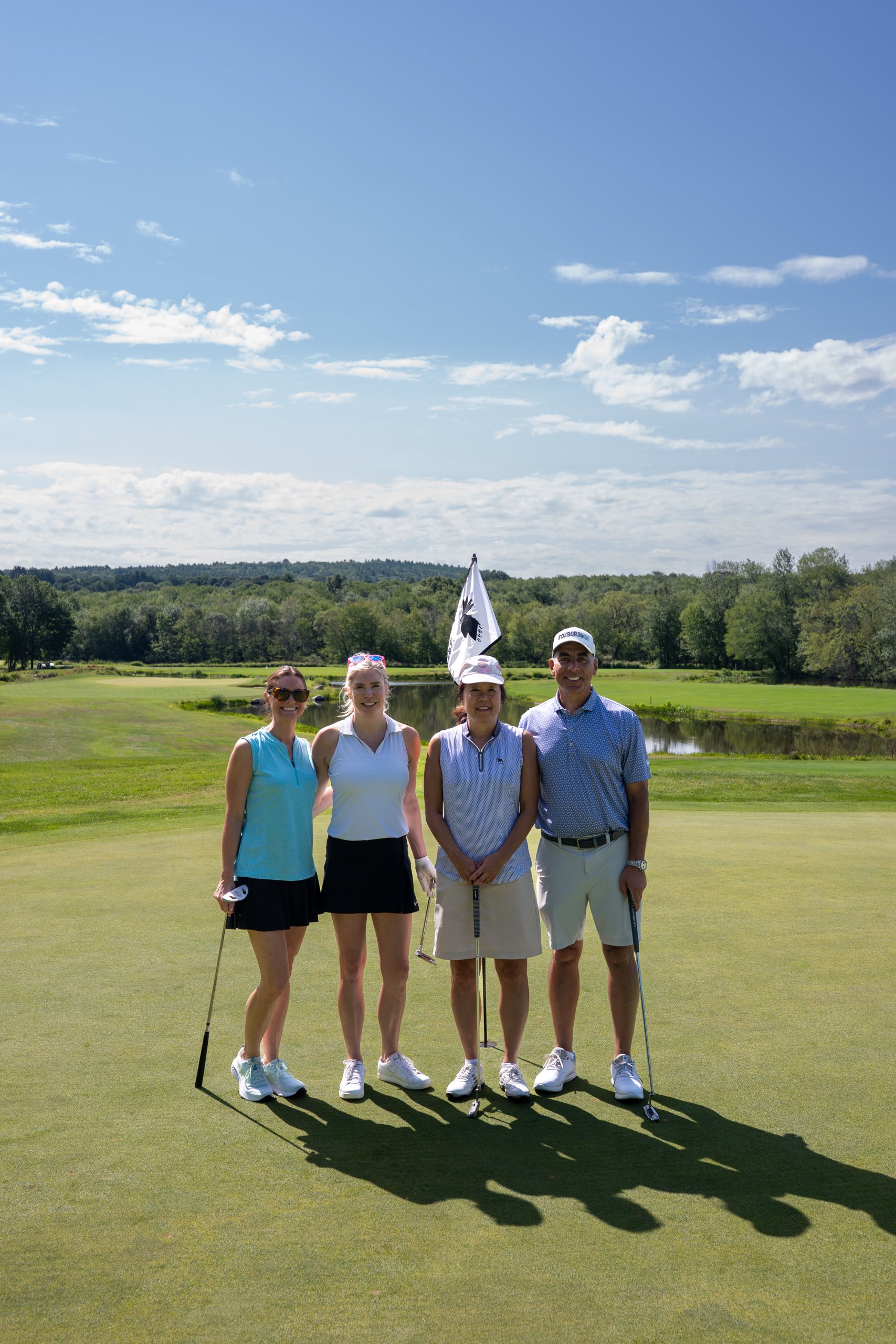 Foursome of three women and a man posing together on the green with their putters, the flag and a pond behind.