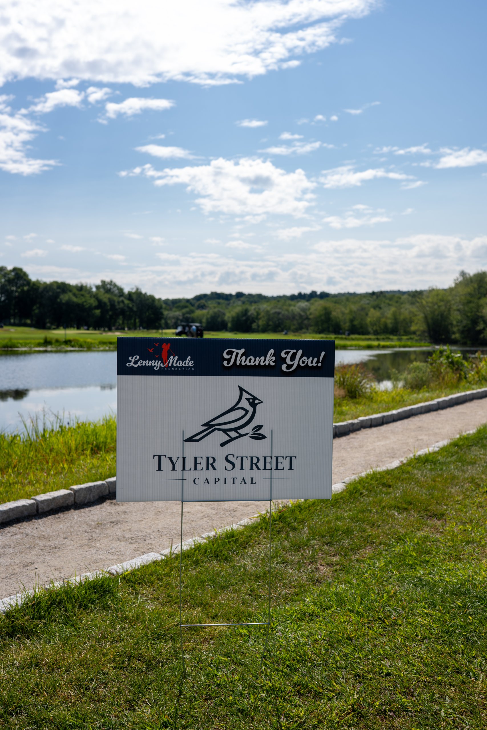 A LennyMade Foundation Thank You sponsor sign for Tyler Street Capital staked beside the pond and cart path.