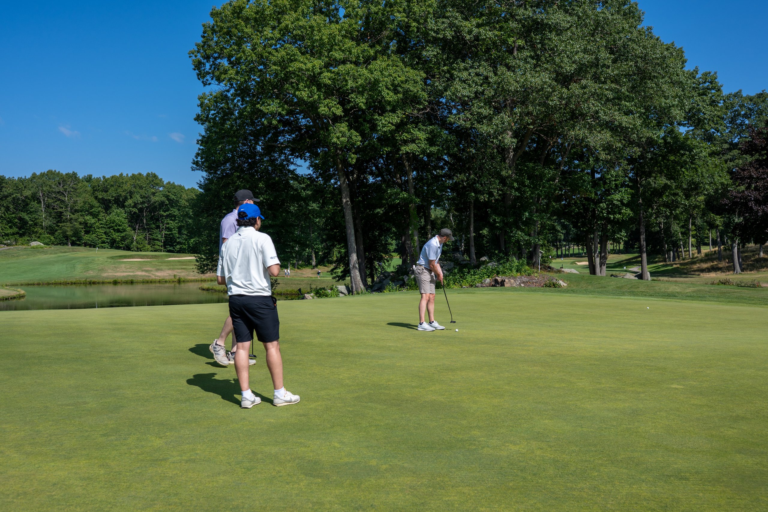 Three golfers on the green, one putting while the others watch, a large shade tree and pond behind.