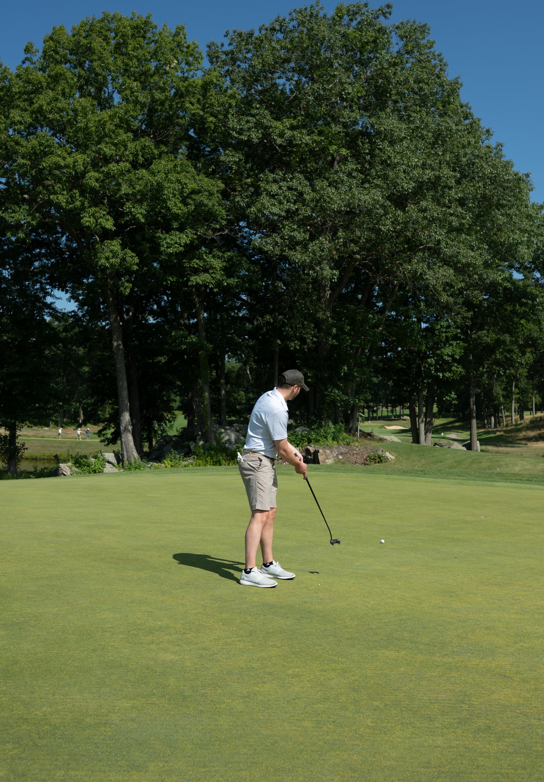 Golfer in a white polo putting on the green, players visible on the fairway across the pond behind him.