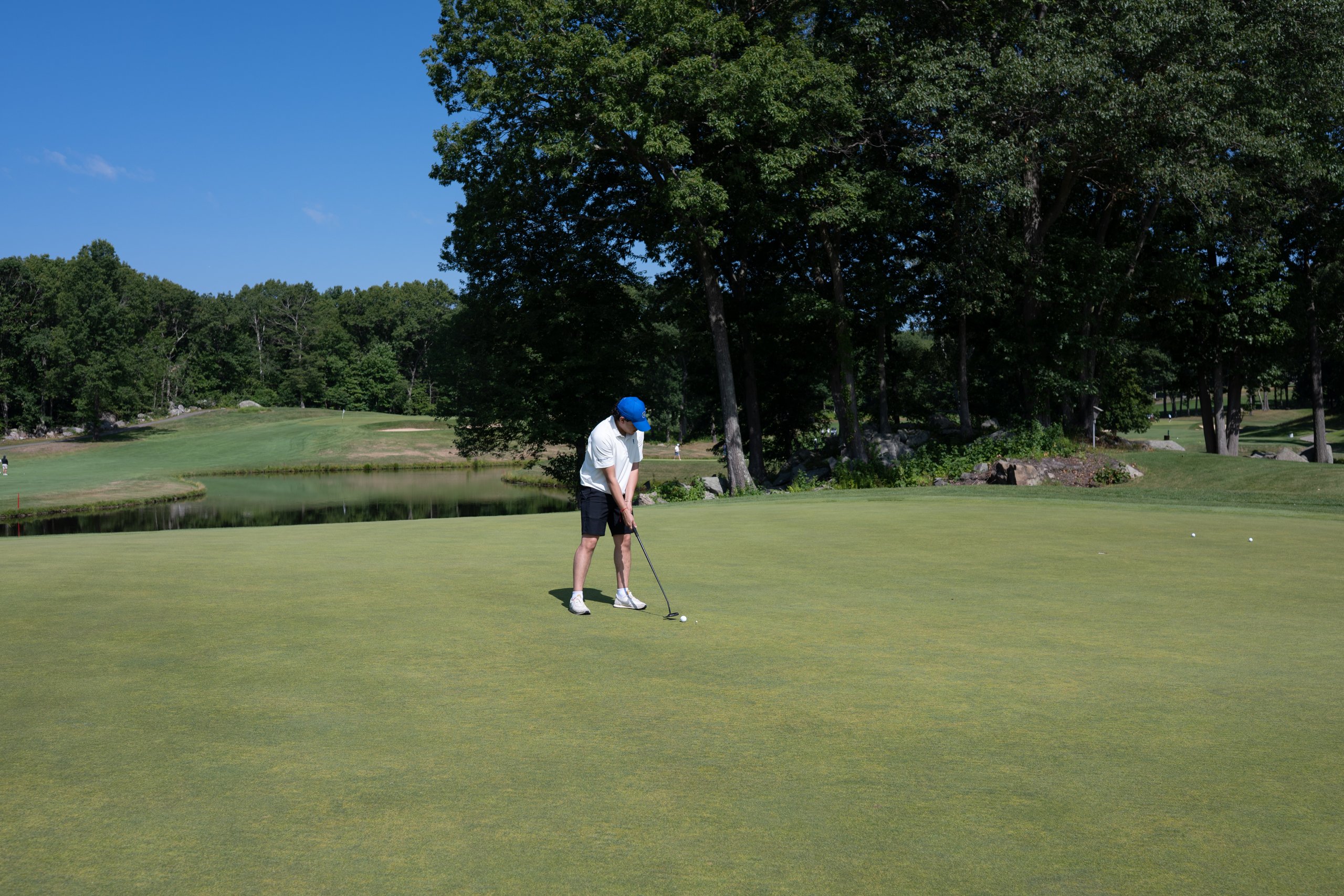 Golfer in a blue cap and white polo putting on the green with a pond and woods in the background.