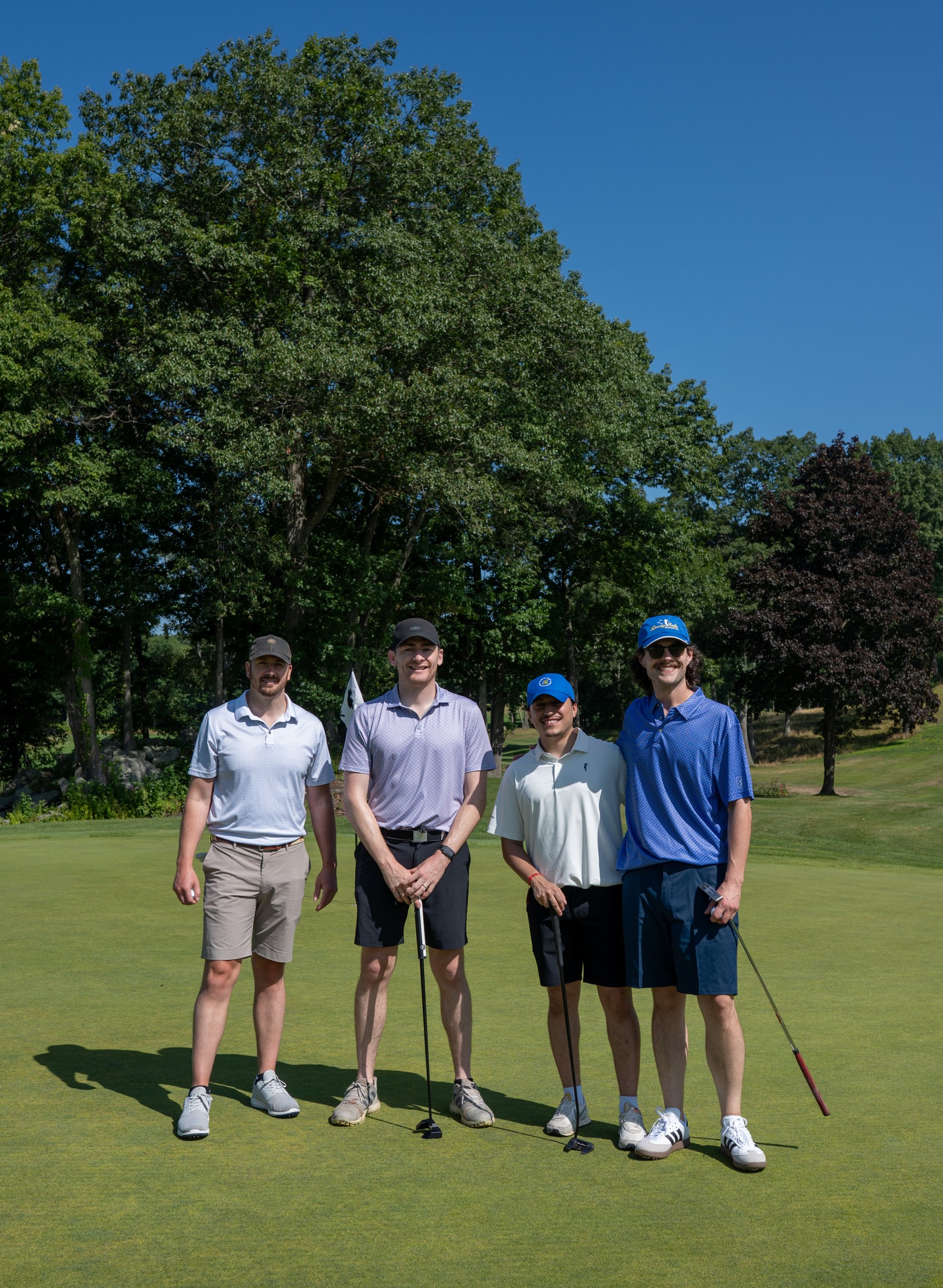 Foursome of four men posing together on the green with their putters, tall trees behind them.