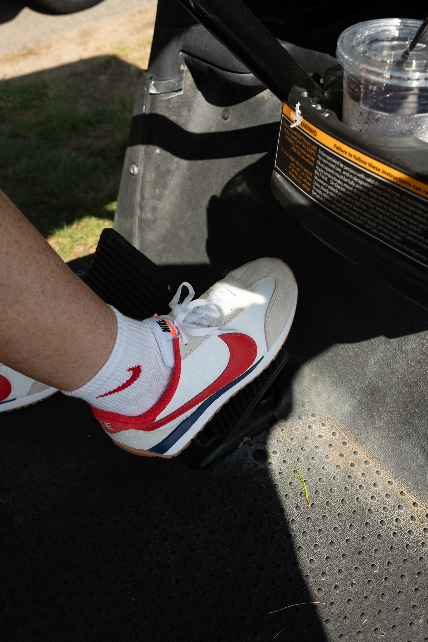 Close-up of a golfer's red-white-and-blue Nike sneaker resting on the cart pedal beside an iced coffee.
