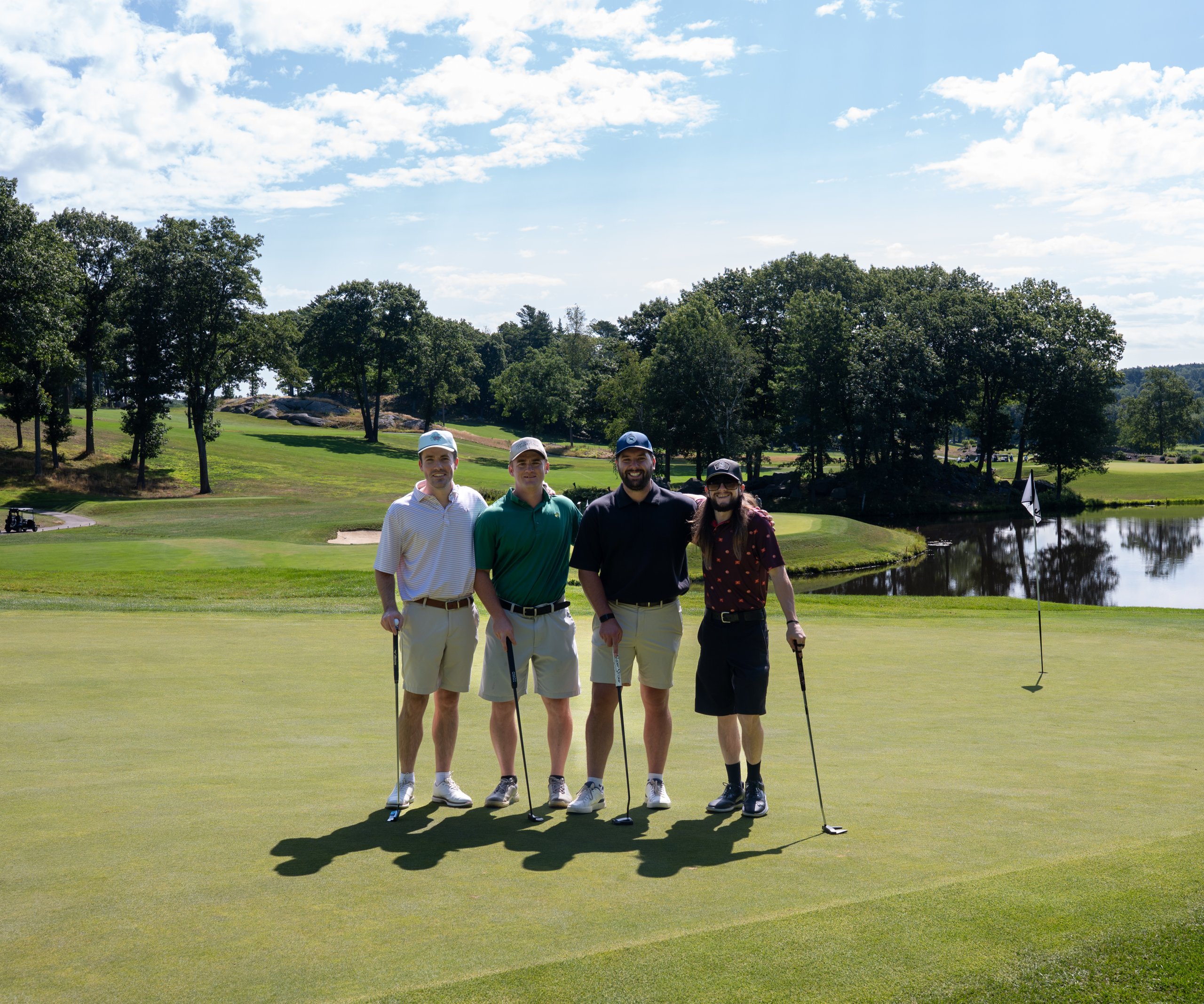 Foursome of four men posing on the green with their putters, a pond and flag behind them.