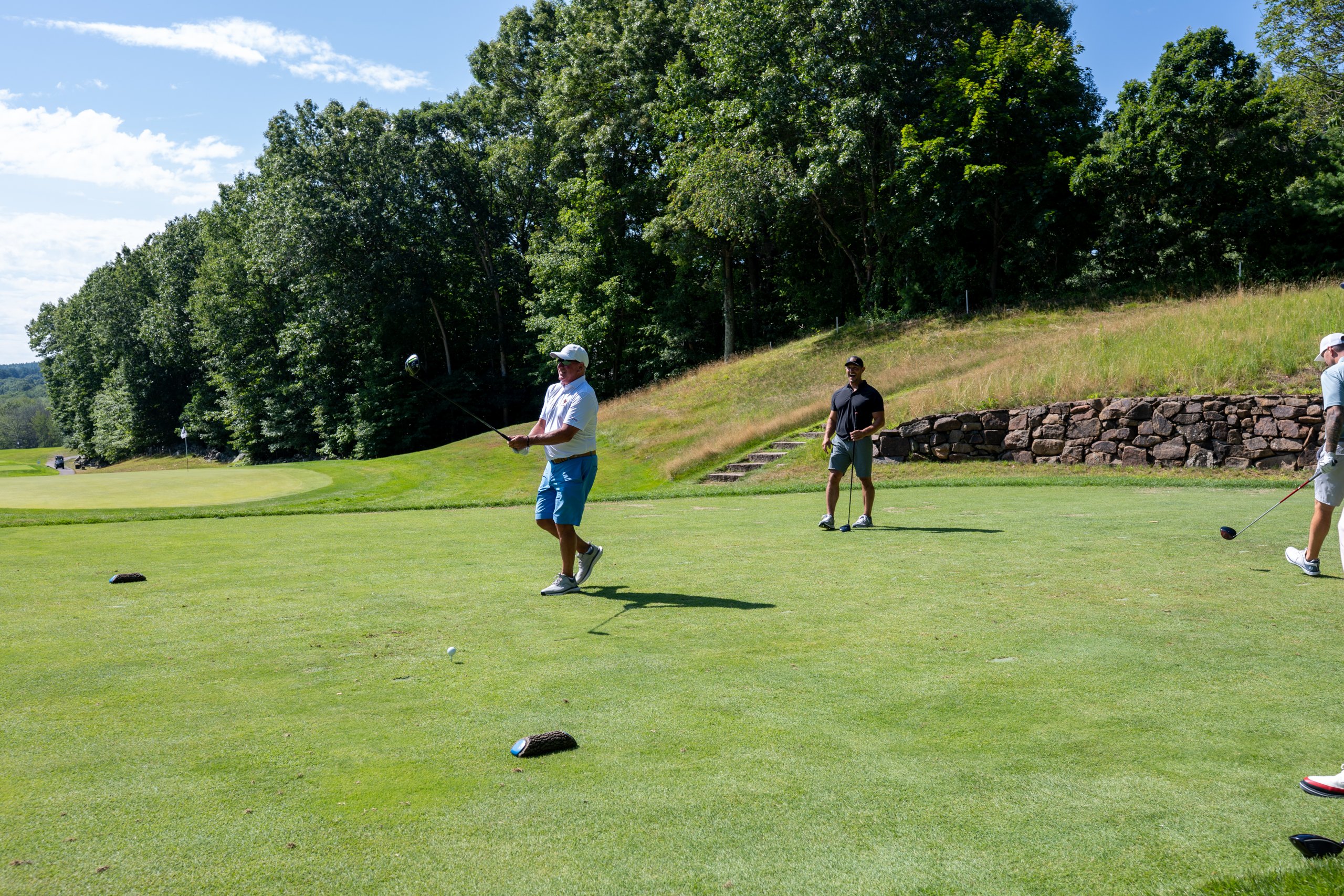 Older golfer in a white polo and blue shorts finishing a tee shot with his driver, a stone retaining wall and grassy bank behind.