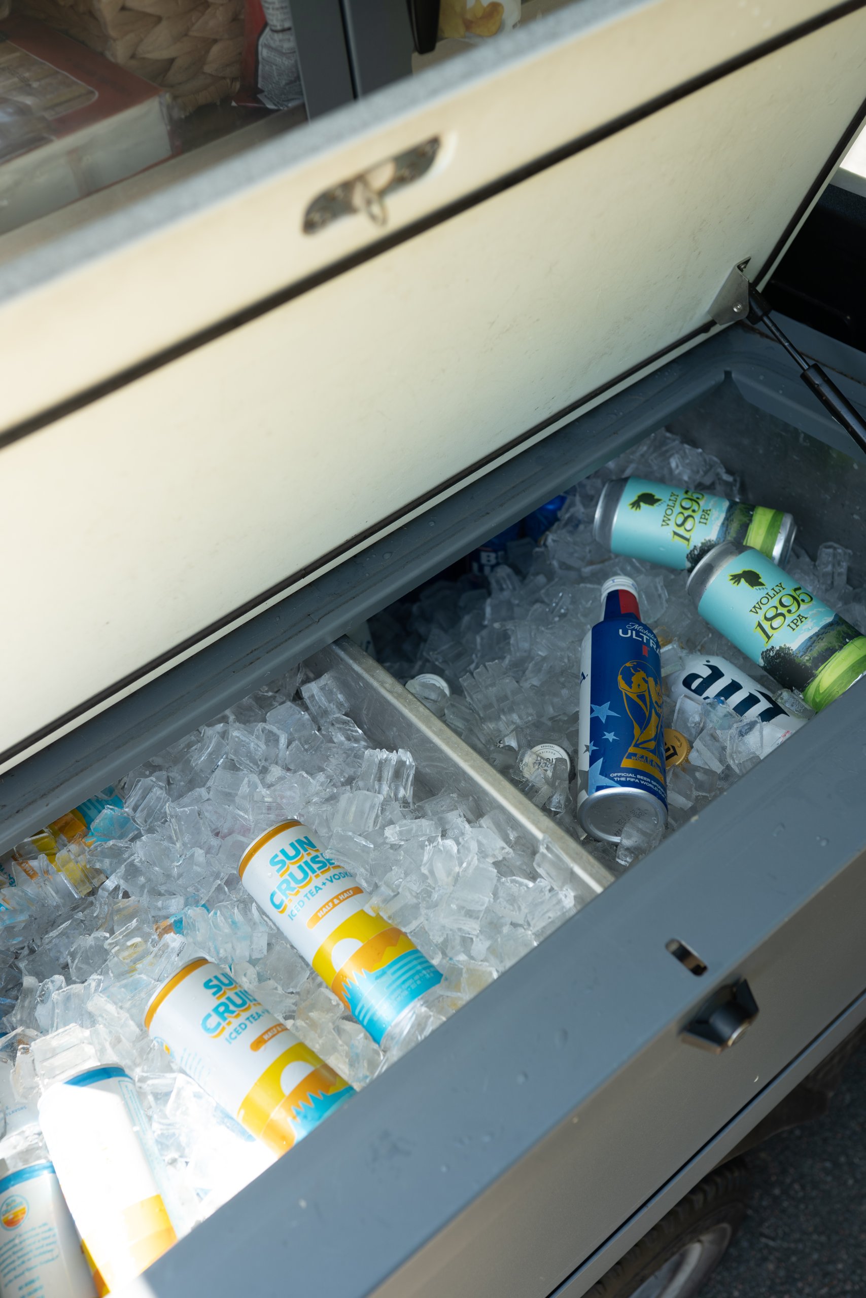 An open golf cart cooler drawer packed with ice and cans of Wolly 1895 IPA, Michelob Ultra, Sun Cruiser and Miller Lite.