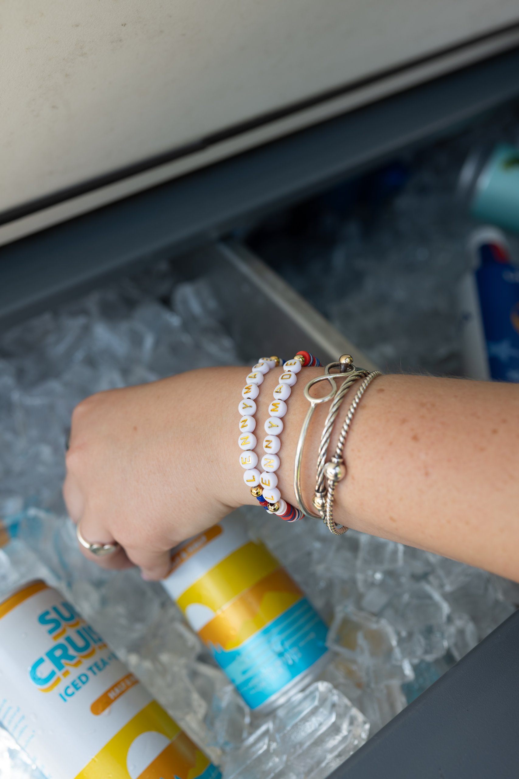 A woman's wrist stacked with beaded LENNYMADE bracelets reaching into the ice-filled cooler for a Sun Cruiser can.