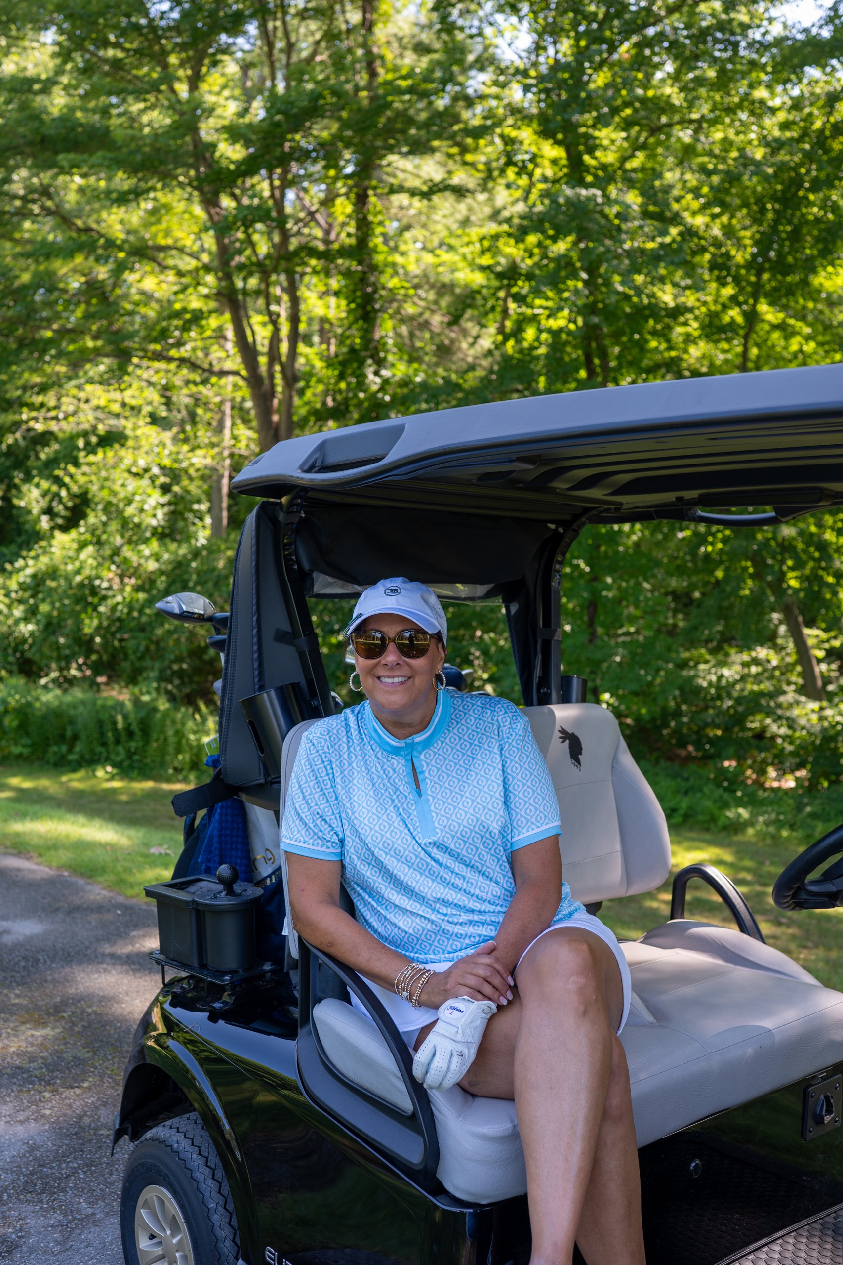 Woman in a light blue polo, white cap and sunglasses sitting on a golf cart and smiling, green trees behind.