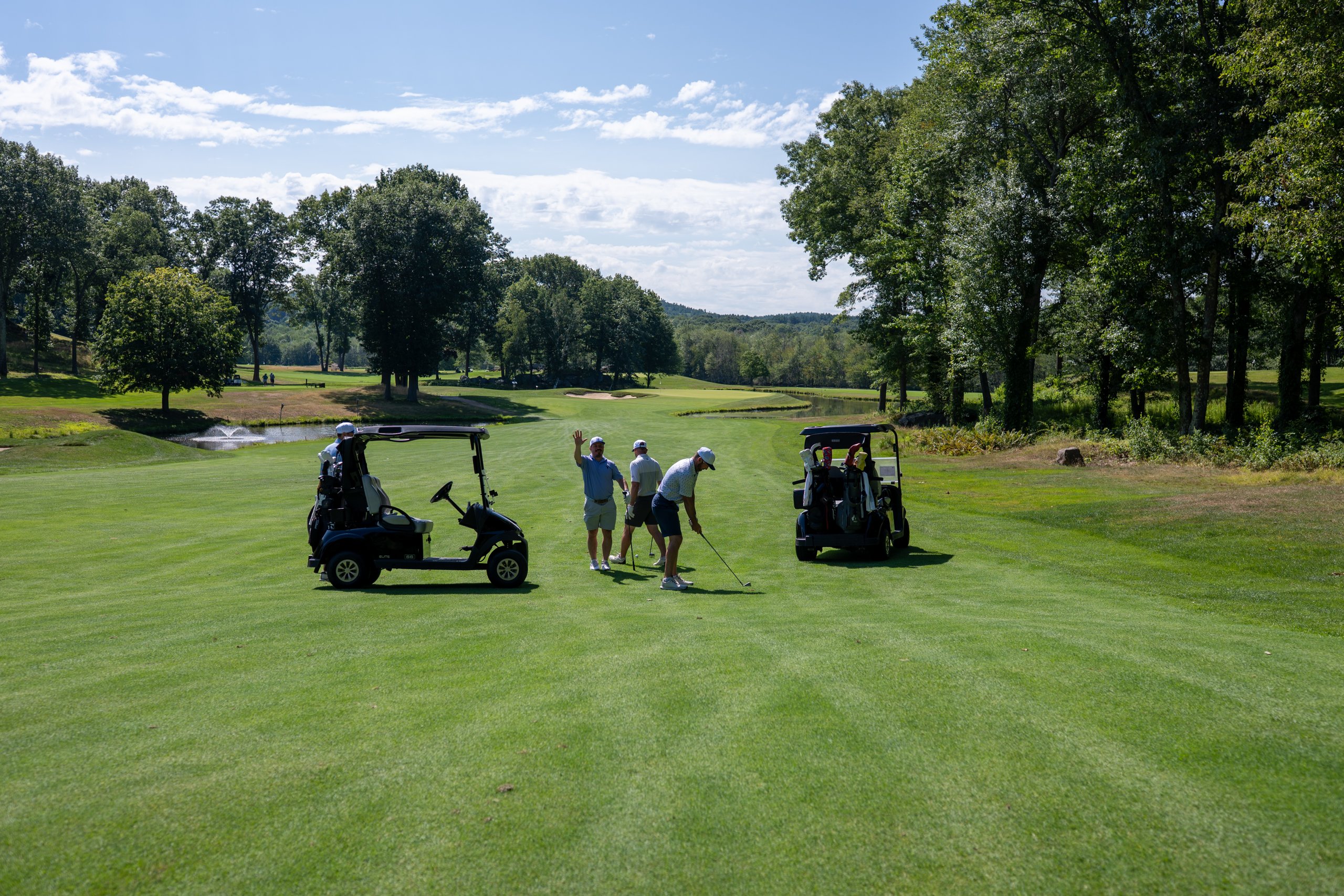 Wide fairway view of a foursome between two carts, one player waving, a fountain pond and green in the distance.