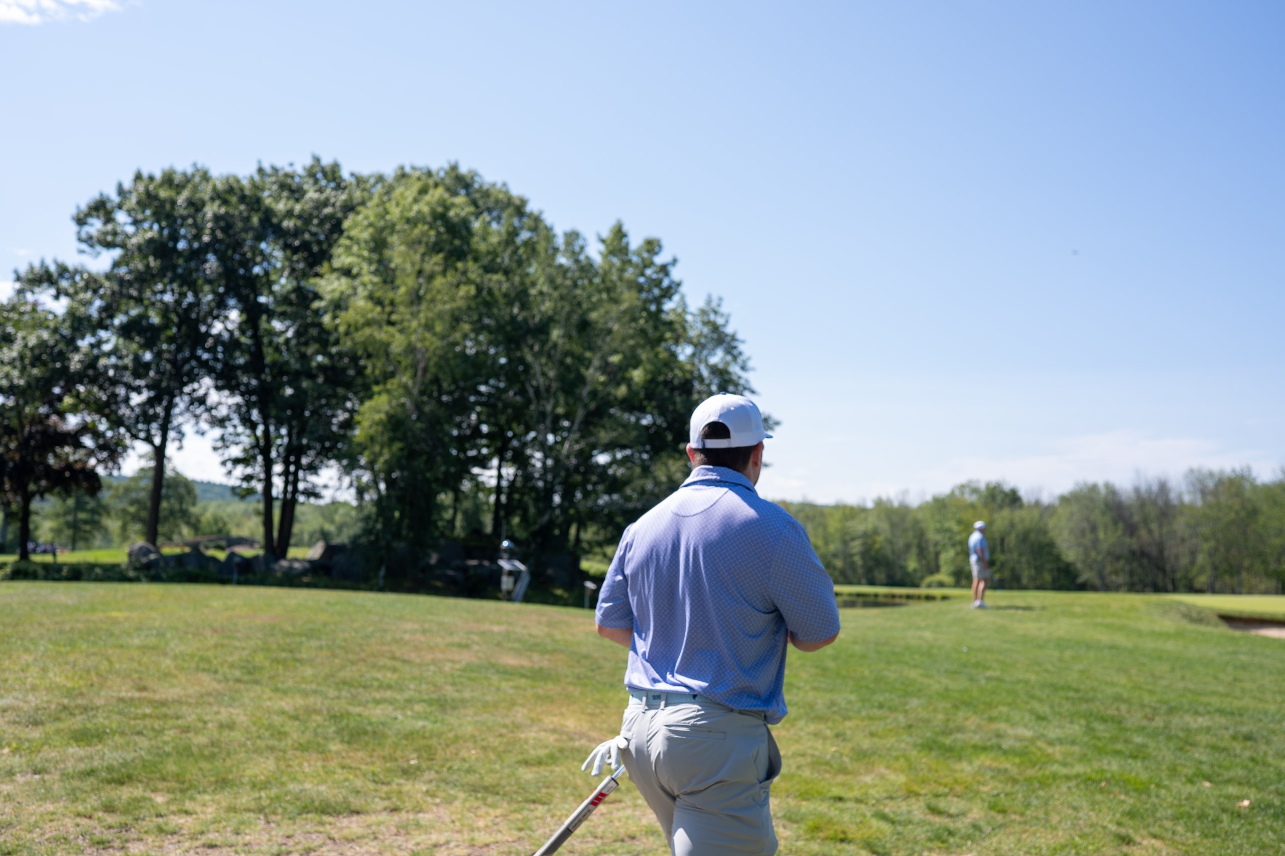 Golfer in a blue polo watching from the fairway as a teammate putts on a distant green beside the pond.