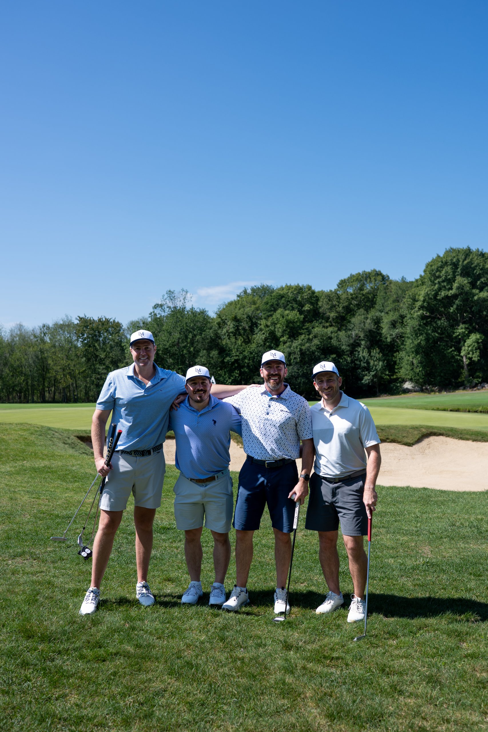 Foursome of four men in matching white caps posing together on the green with their putters, a bunker behind.