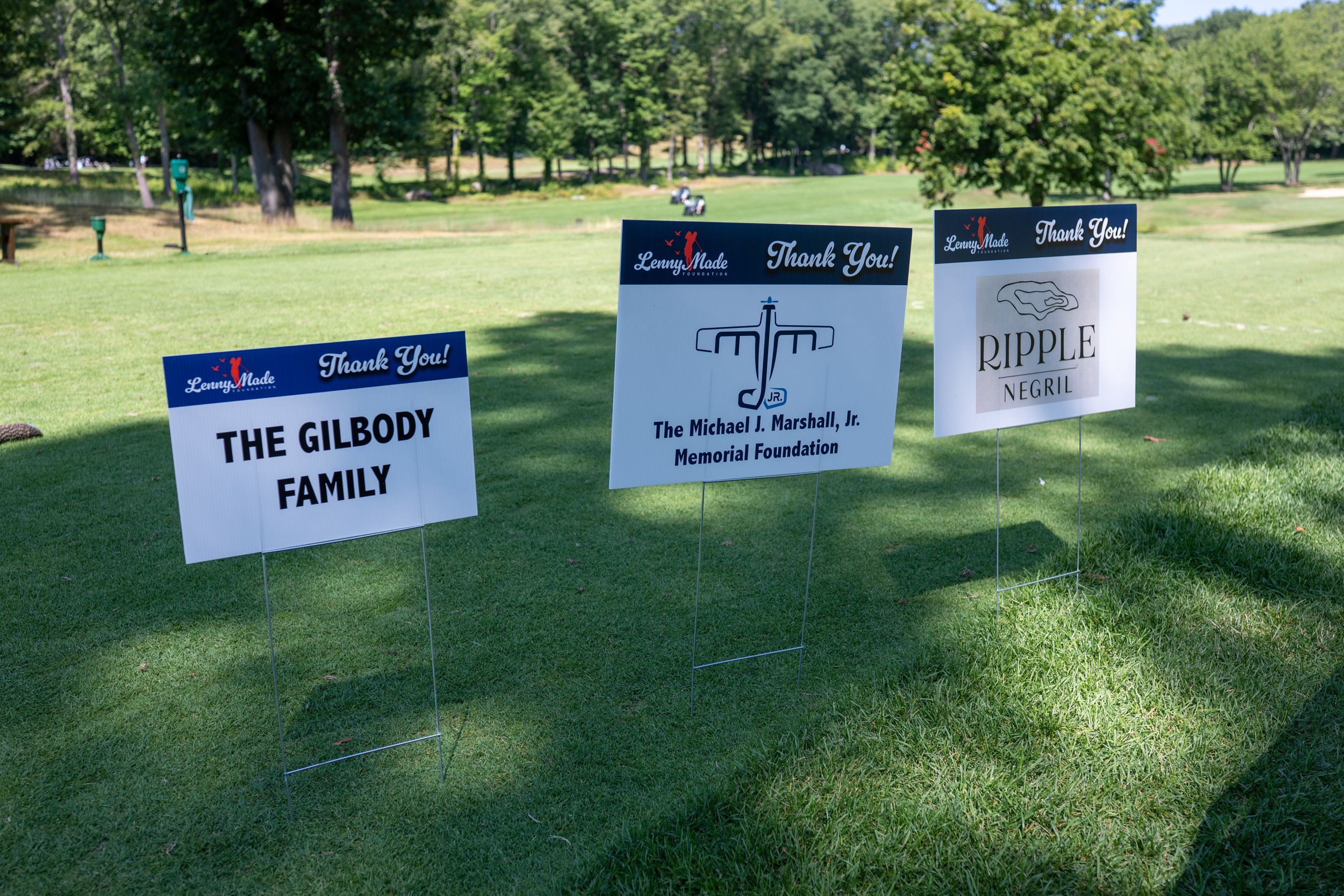 Three LennyMade Foundation Thank You sponsor signs staked together on the grass: the Gilbody Family, the Michael J. Marshall Jr. Memorial Foundation and Ripple Negril.