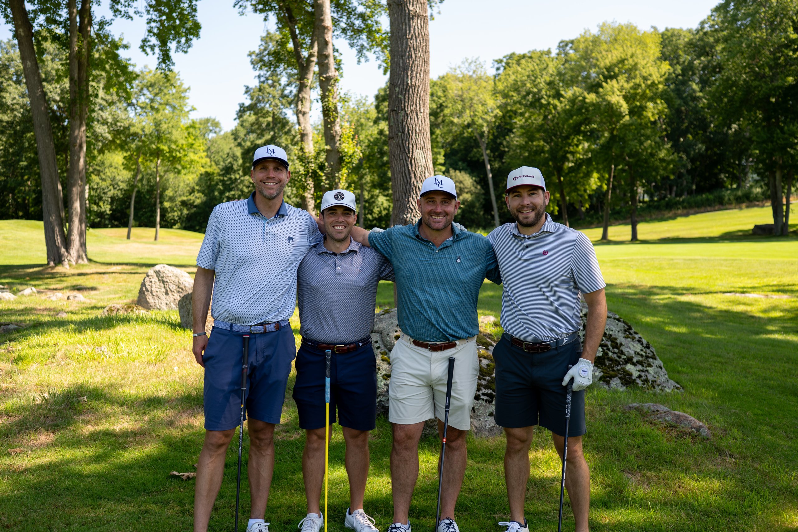 Foursome of four men in matching caps posing together on the grass with their putters, a mossy boulder behind.