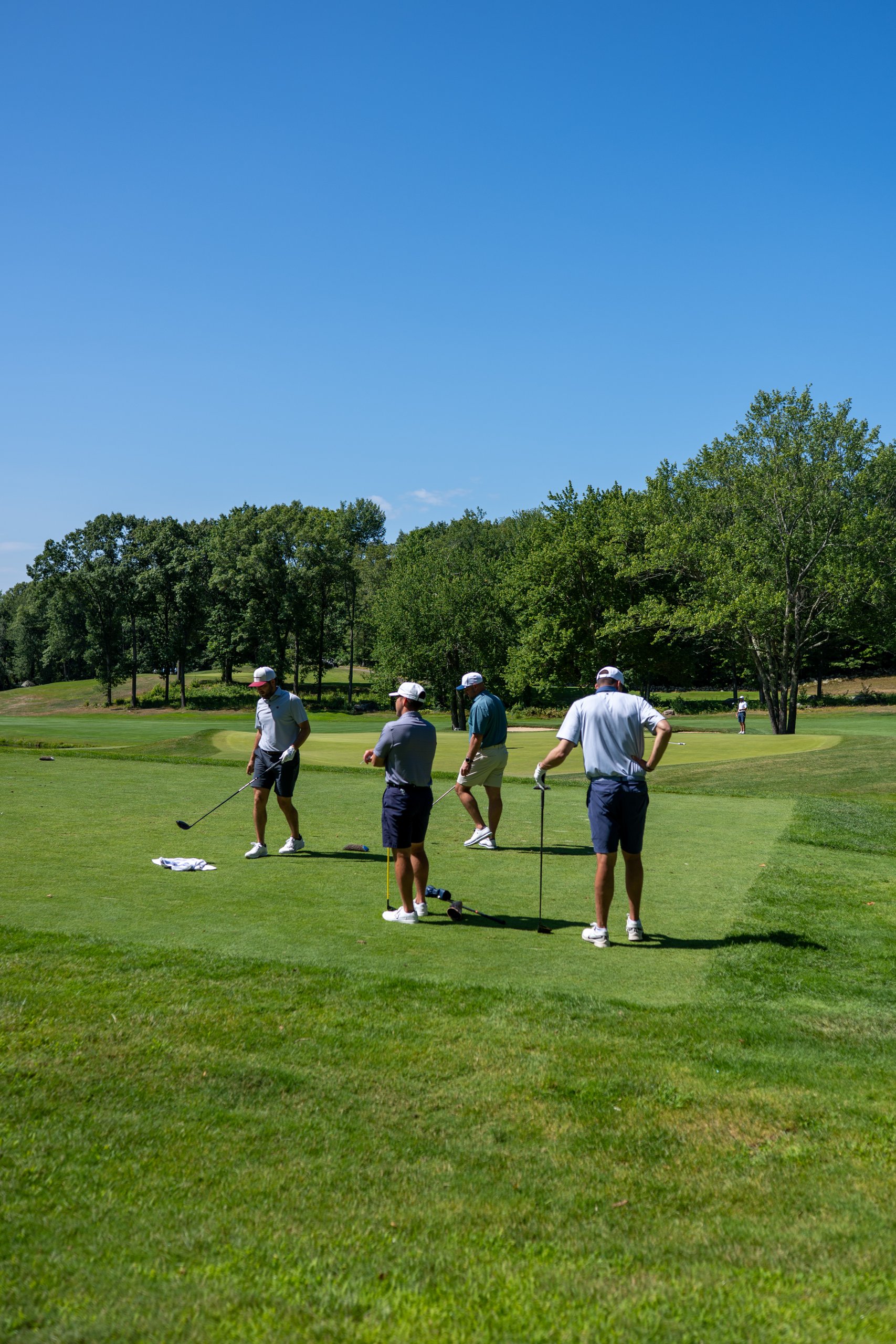 Four golfers on the tee, one raising a hand, with the wooded course stretching out behind them.