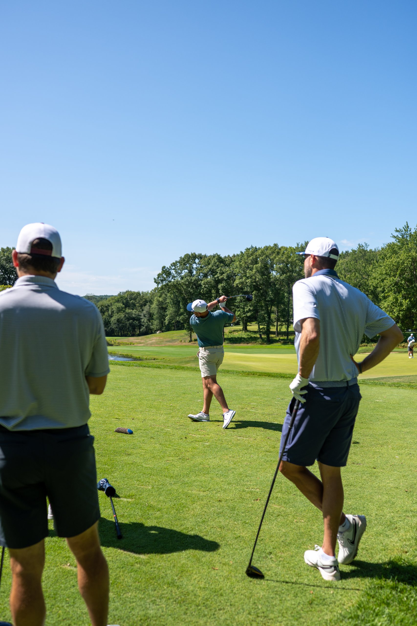 Two golfers watching from the tee as a teammate drives, a pond and green in the distance.