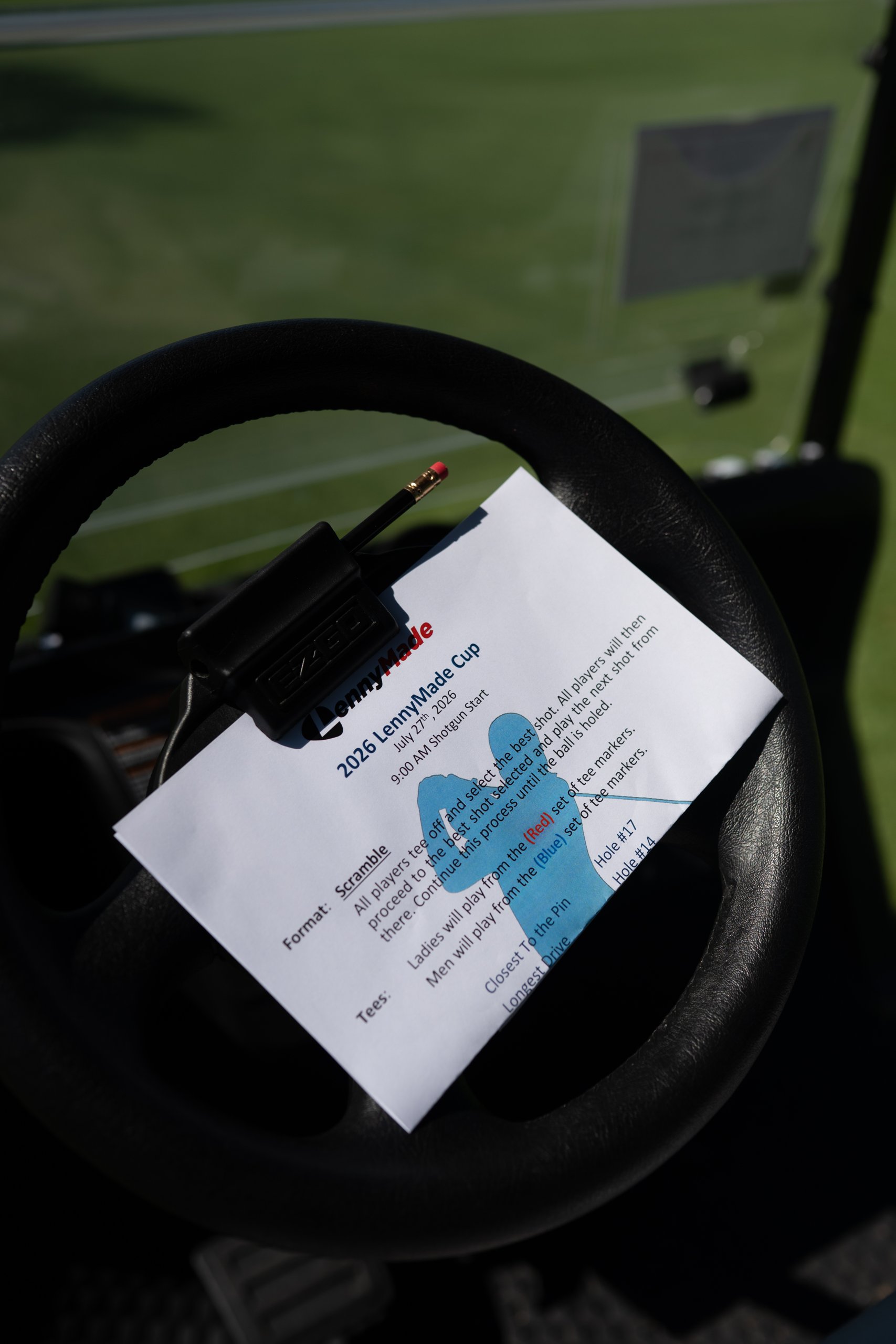 Close-up of the 2026 LennyMade Cup rules sheet clipped to a cart steering wheel, listing the July 27 9 AM shotgun scramble format.
