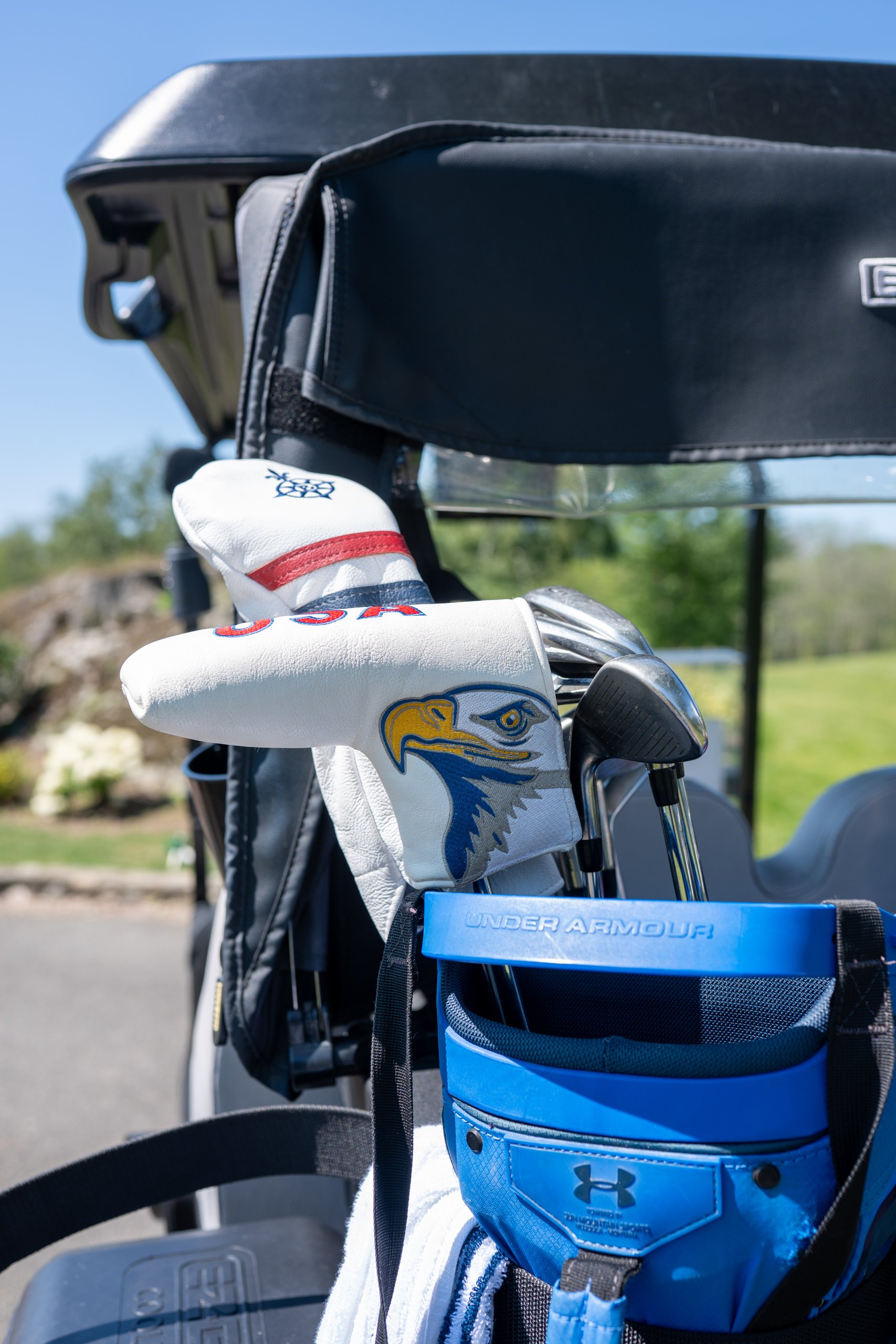 A USA driver headcover and an eagle-head club cover on an Under Armour golf bag mounted on the back of a cart.