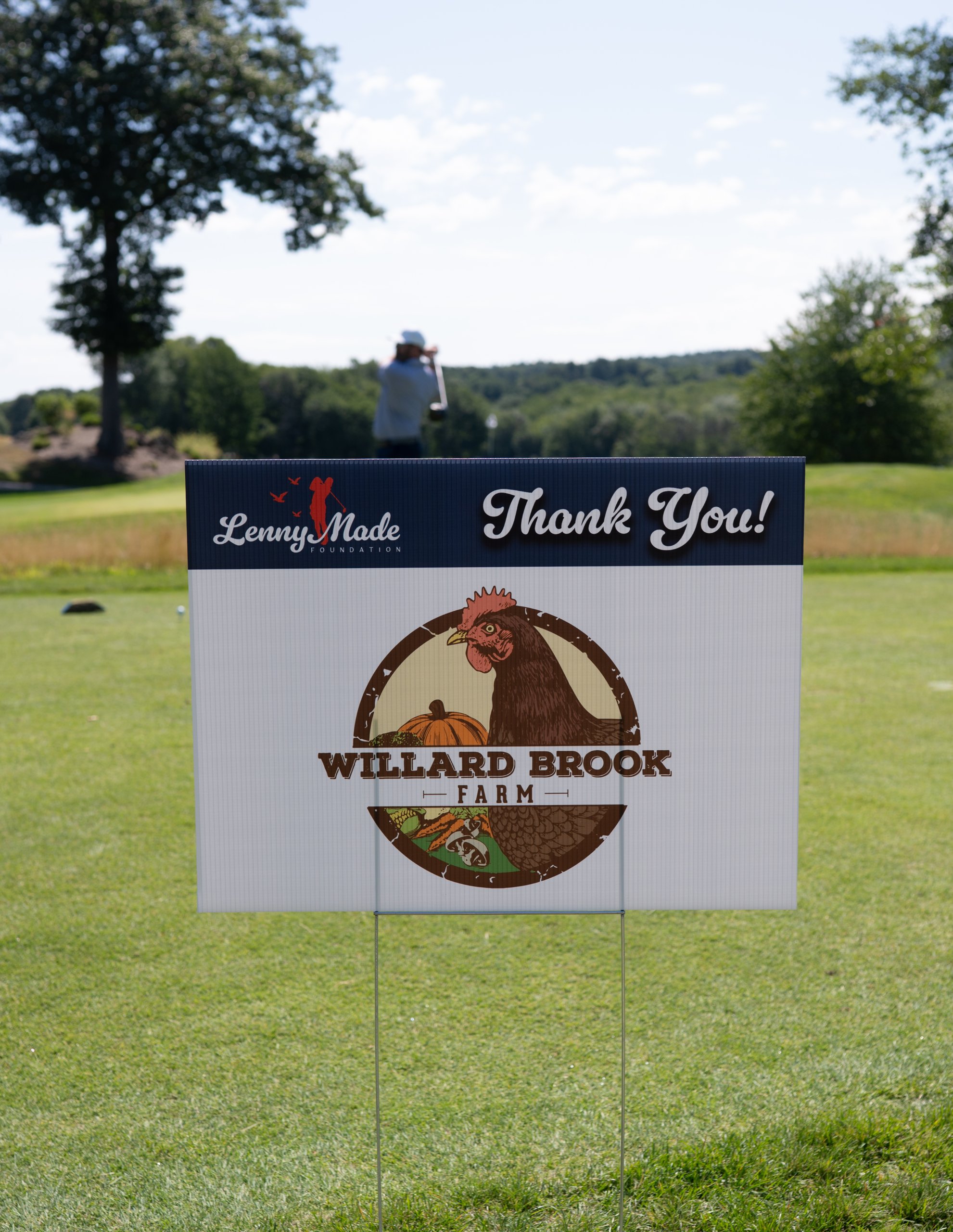A LennyMade Foundation Thank You sponsor sign for Willard Brook Farm, with its rooster logo, on the course.