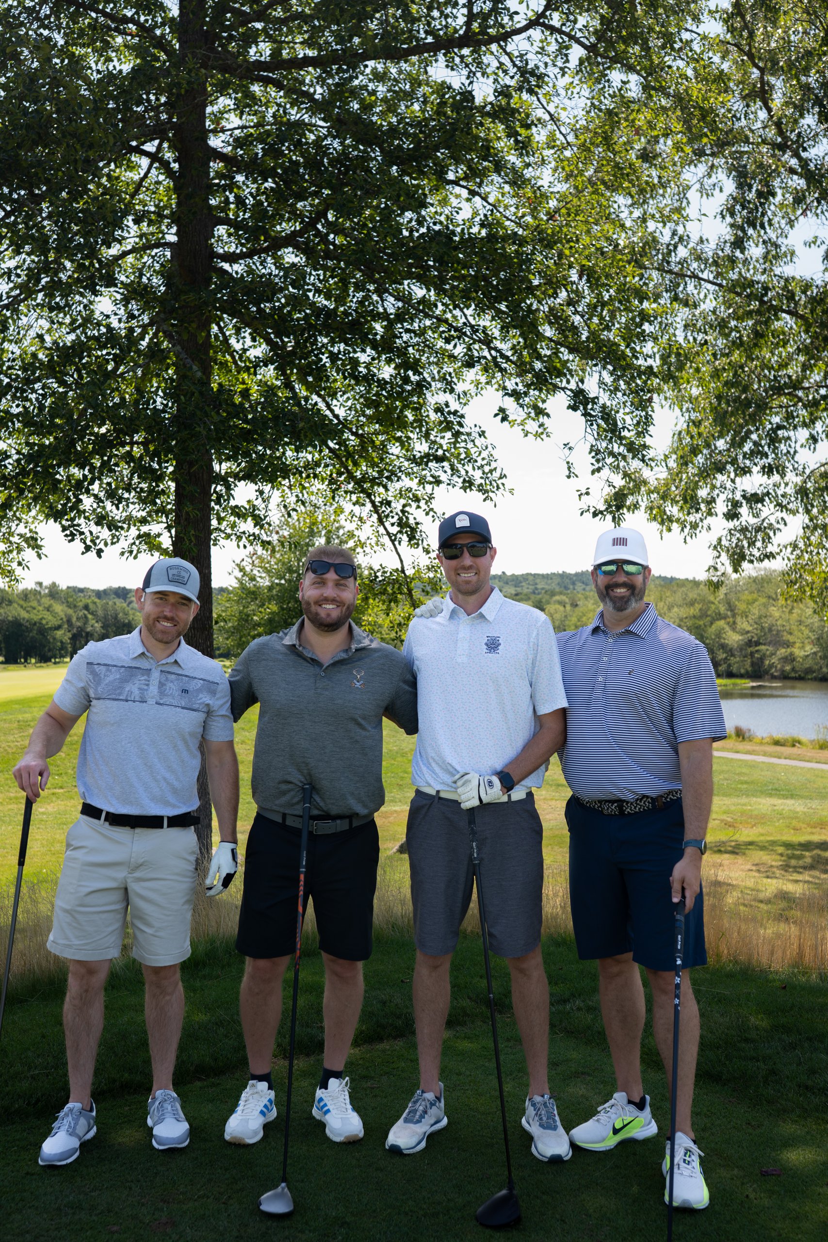 Foursome of four men posing together on the tee beneath a shade tree, a pond and treeline behind them.
