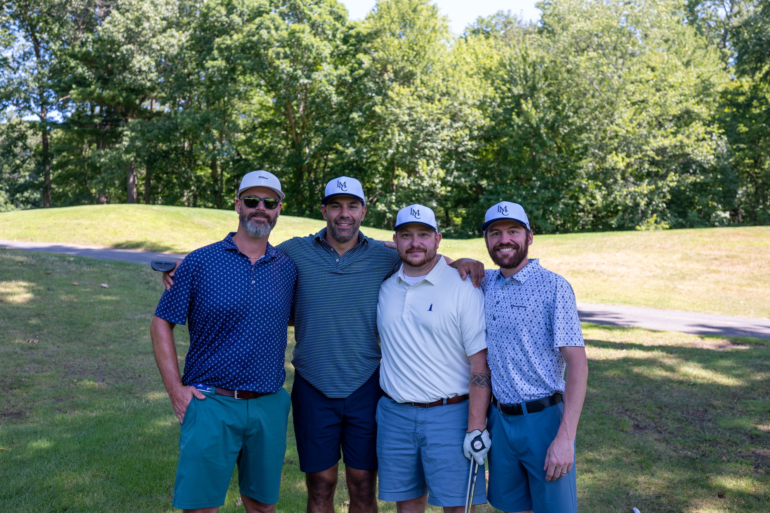 Foursome of four men in matching blue LMC caps posing arm in arm on the cart path.