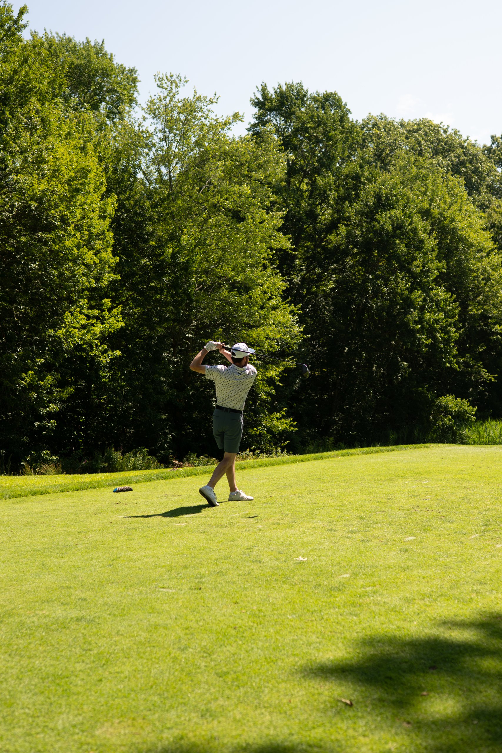 A lone golfer finishing his tee-shot follow-through on the fairway, tall trees filling the background.