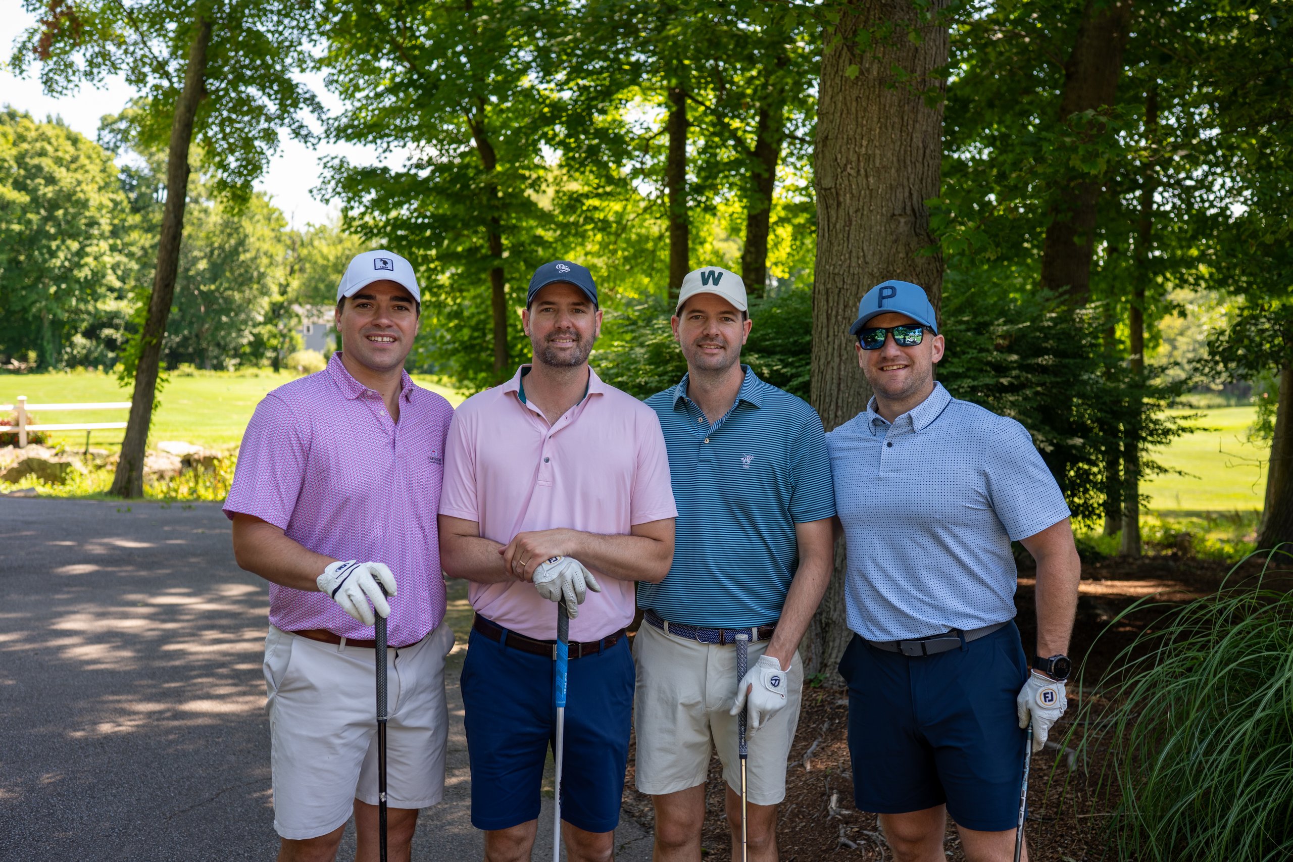 Foursome of four men posing with their clubs on the shaded cart path, the fairway and pond behind.