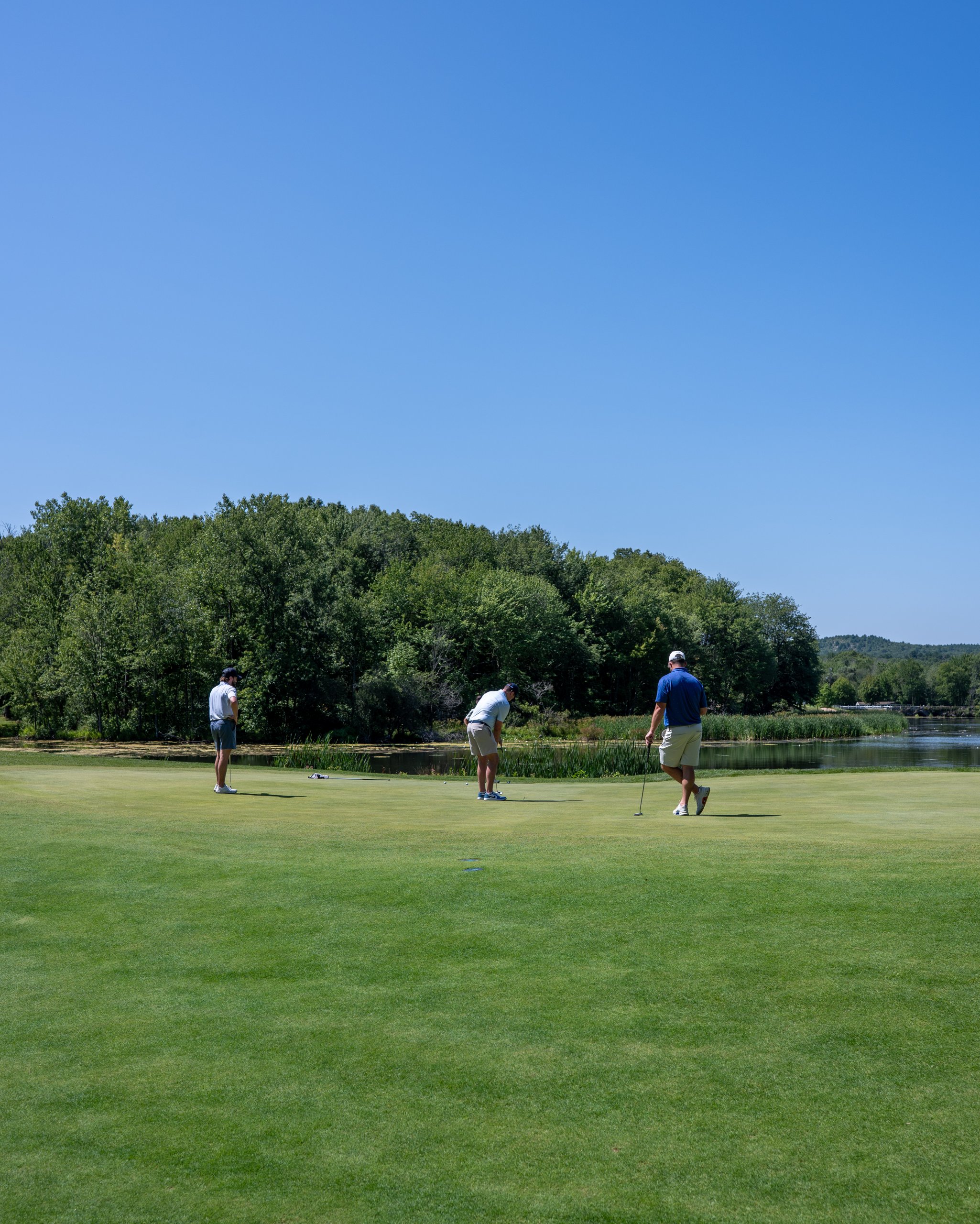 Three golfers putting out on a green beside a lily-pad pond, a wide view of the wooded course behind.