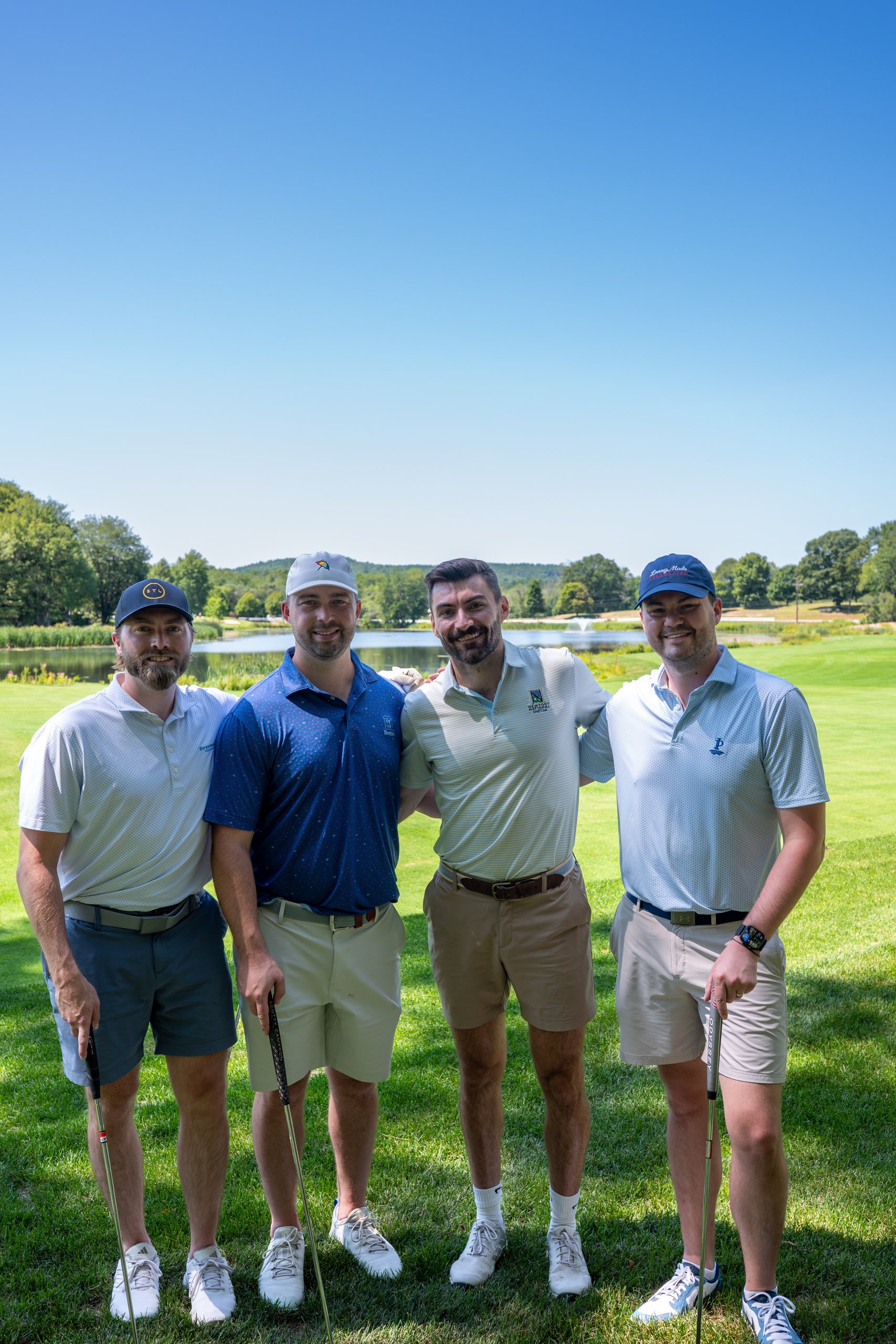 Foursome of four men posing together on the green with their putters, a fountain pond behind them.