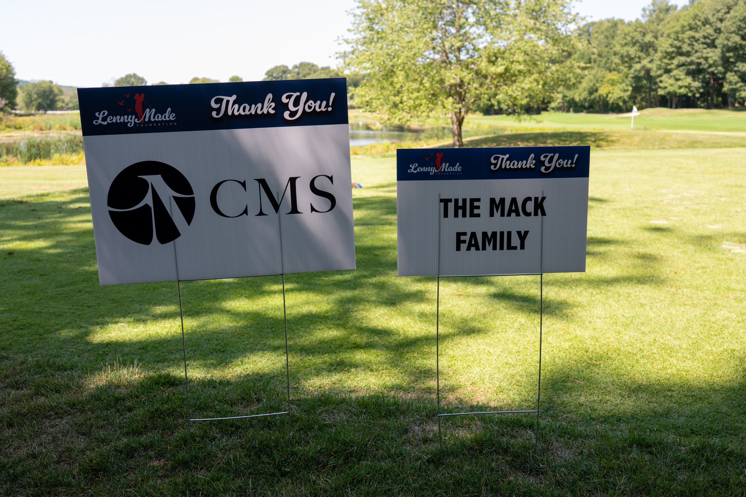 Two LennyMade Foundation sponsor signs side by side on the fairway: CMS and The Mack Family.