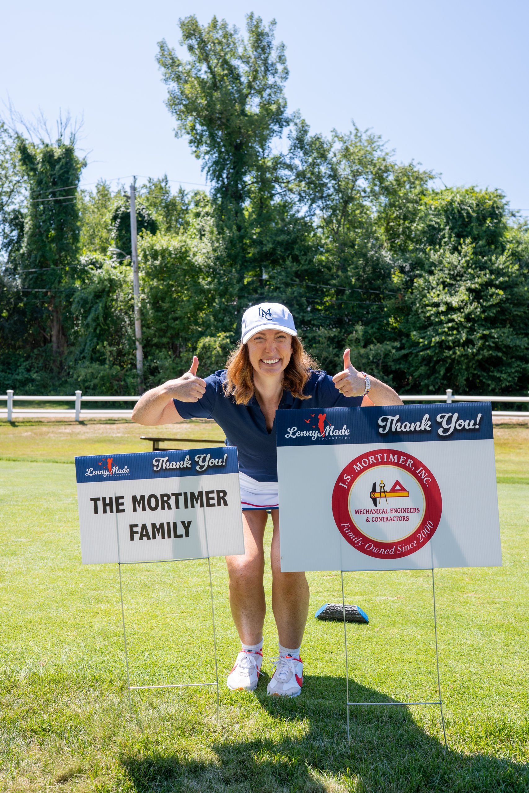 A volunteer in an LMC cap giving two thumbs up between the Mortimer Family and J.S. Mortimer sponsor signs.